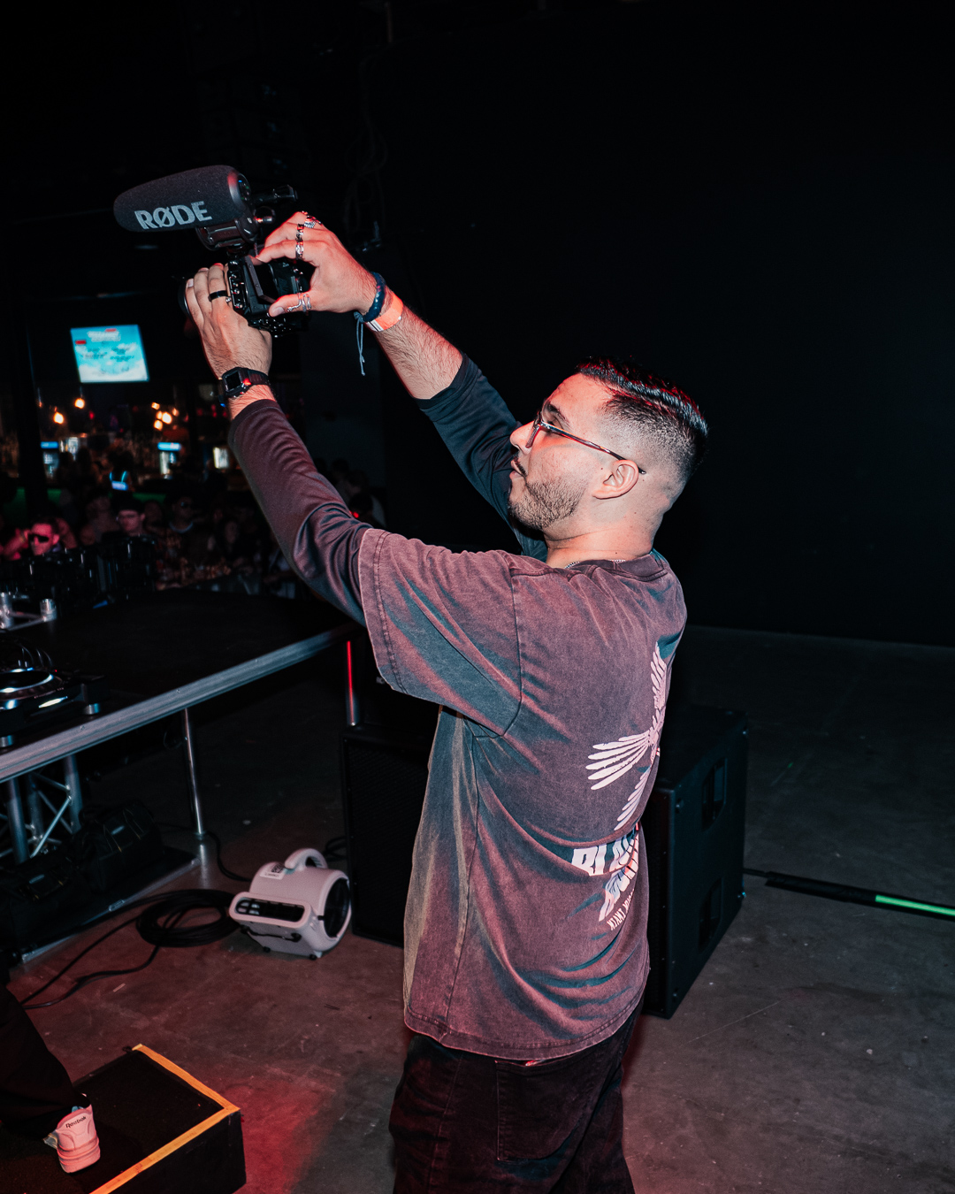 Person holding a camera on stage with a dark background, wearing glasses and a long-sleeve shirt, facing right.