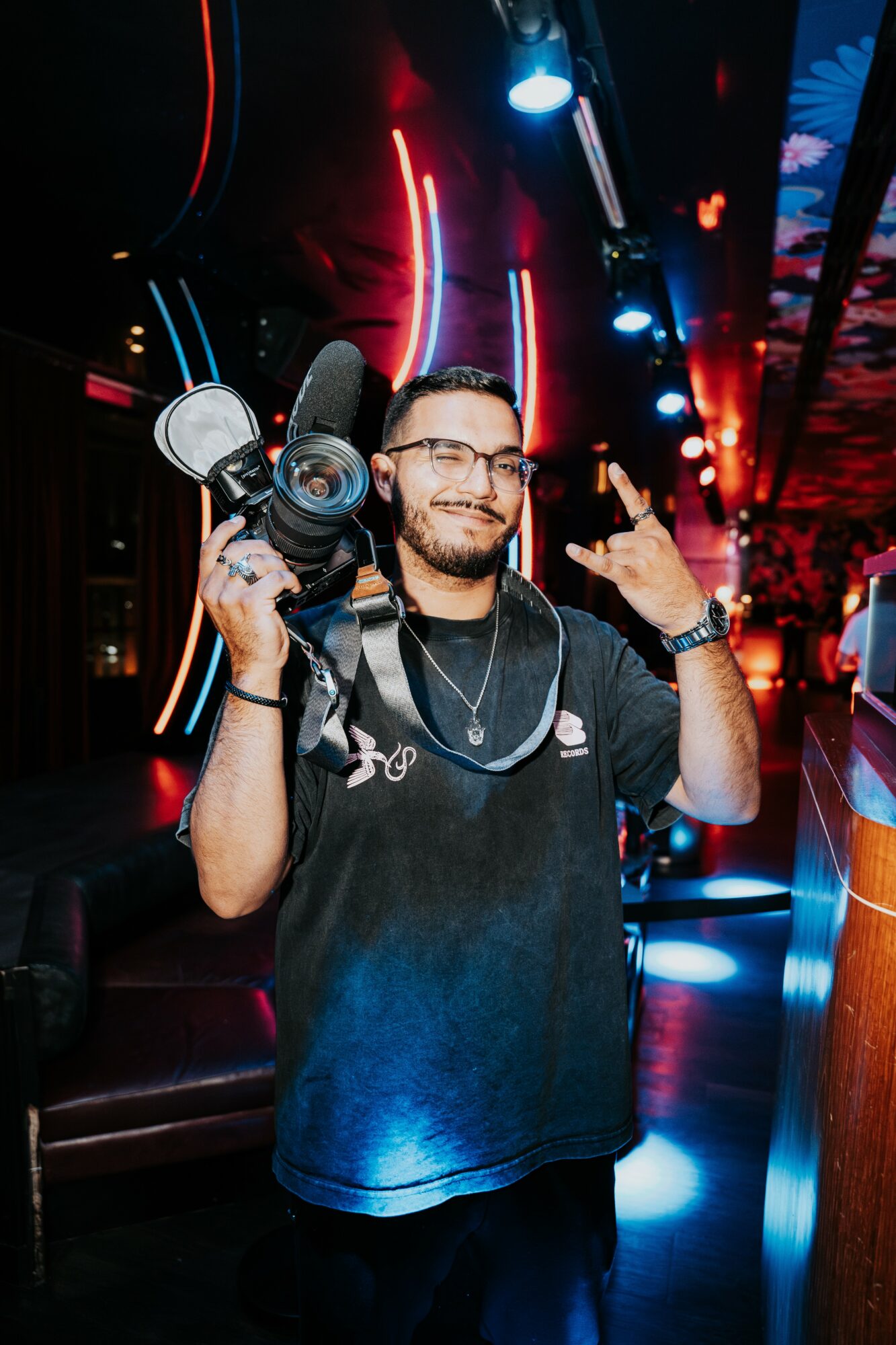 Man holding professional video camera in a colorful, neon-lit indoor setting, making a hand gesture, smiling.
