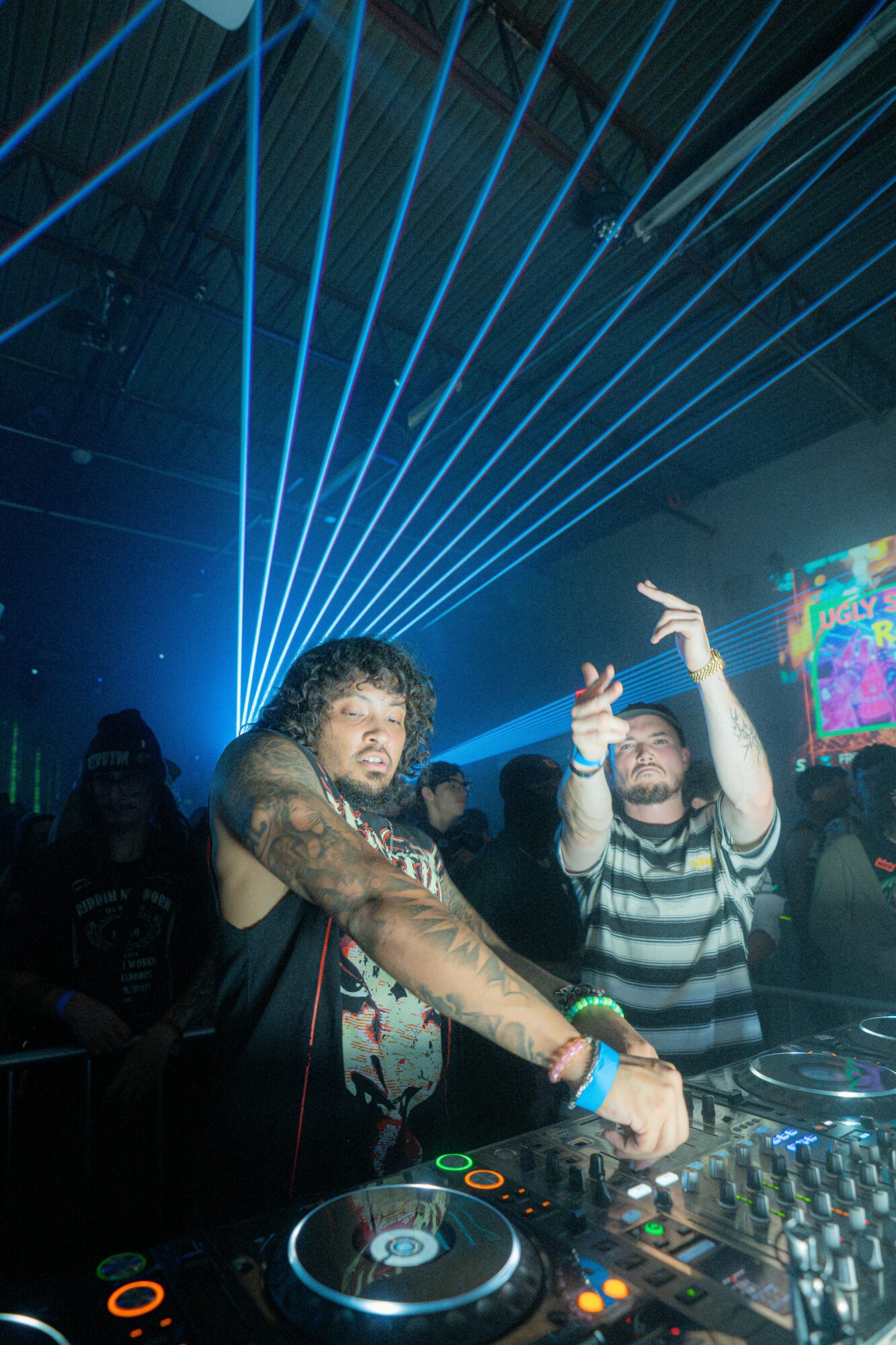 Two DJs perform with laser lights and a crowd in a nightclub setting, one DJ adjusting equipment, the other raising hands.