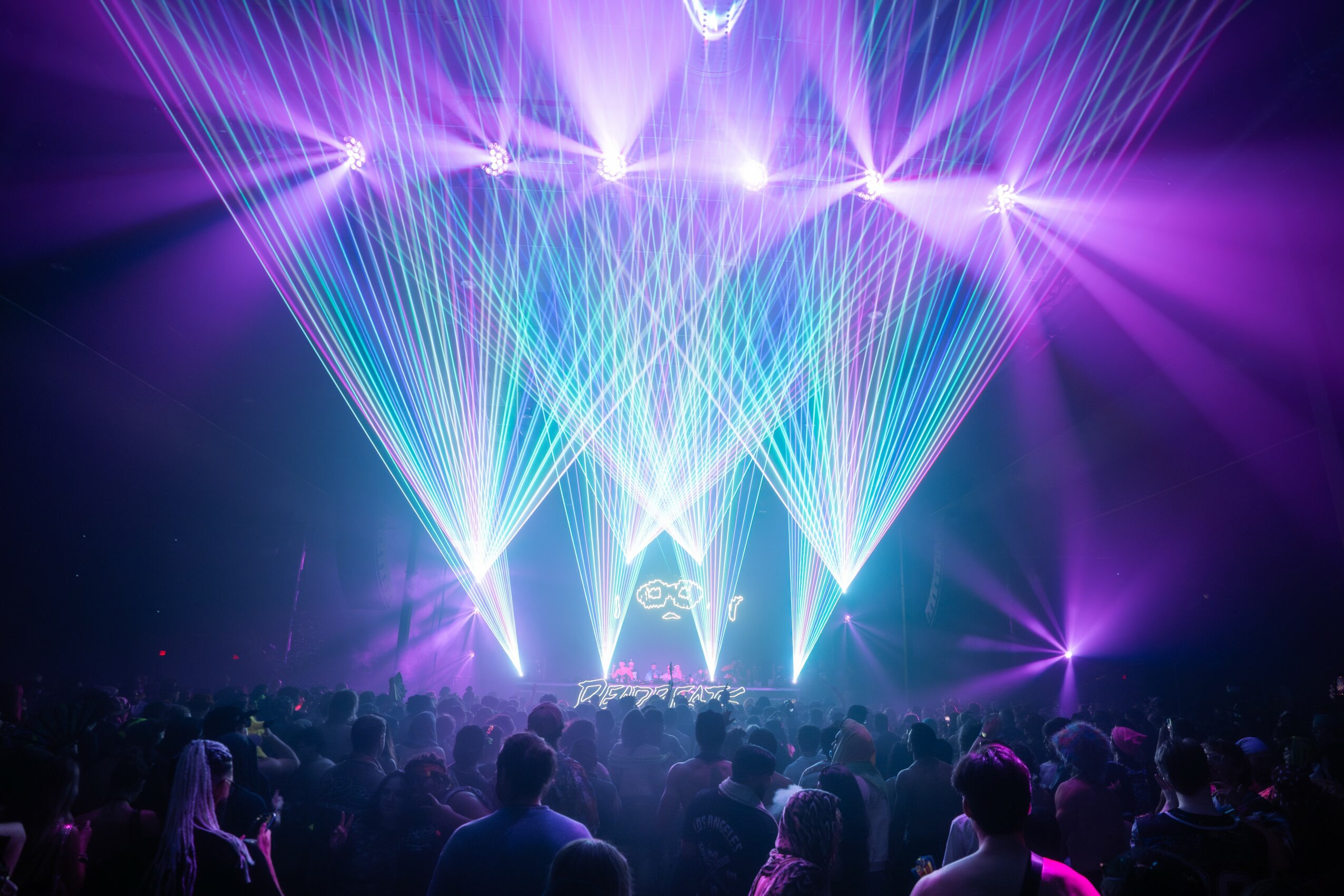 Concert scene with colorful laser lights and a crowd of people watching the stage, illuminated by purple and blue lights.
