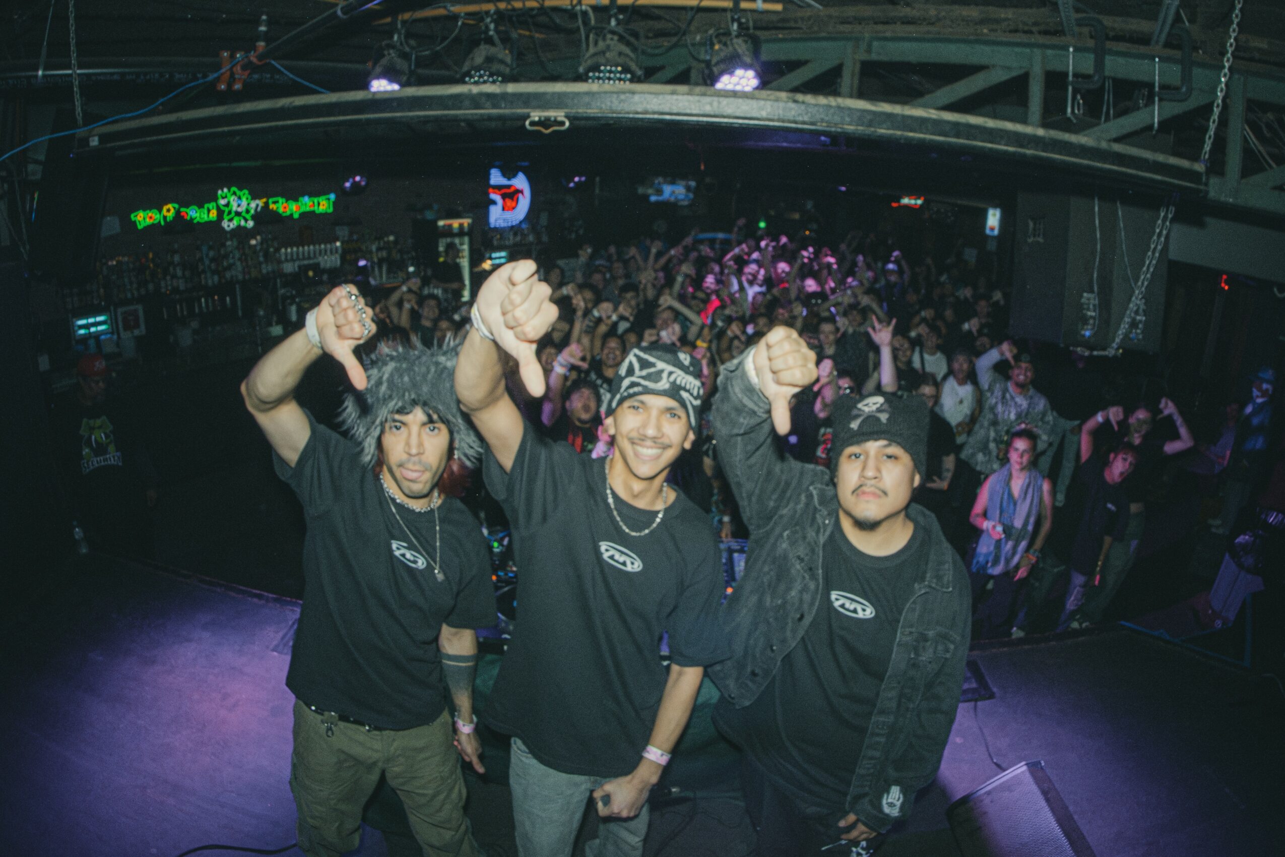 Three young men in black shirts and beanies pose with fists raised in a crowded indoor event space, with a large audience in the background.