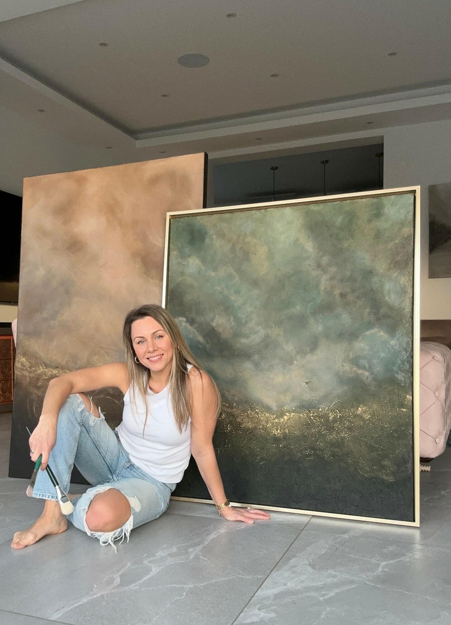 Young woman sitting on the floor next to two large abstract paintings, smiling, in an indoor setting.