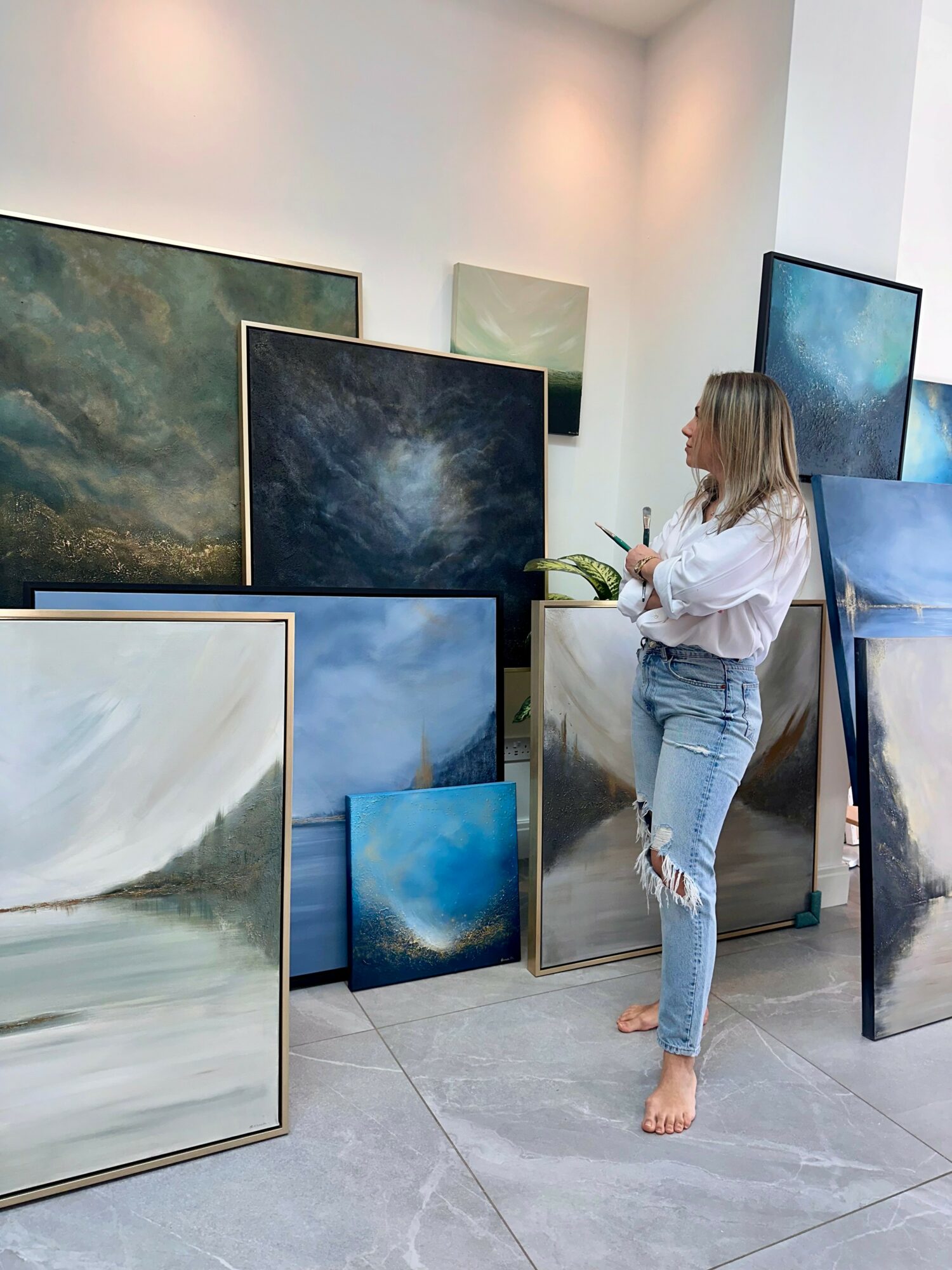 Woman with blonde hair, white shirt, and ripped jeans stands barefoot among paintings in an art gallery.