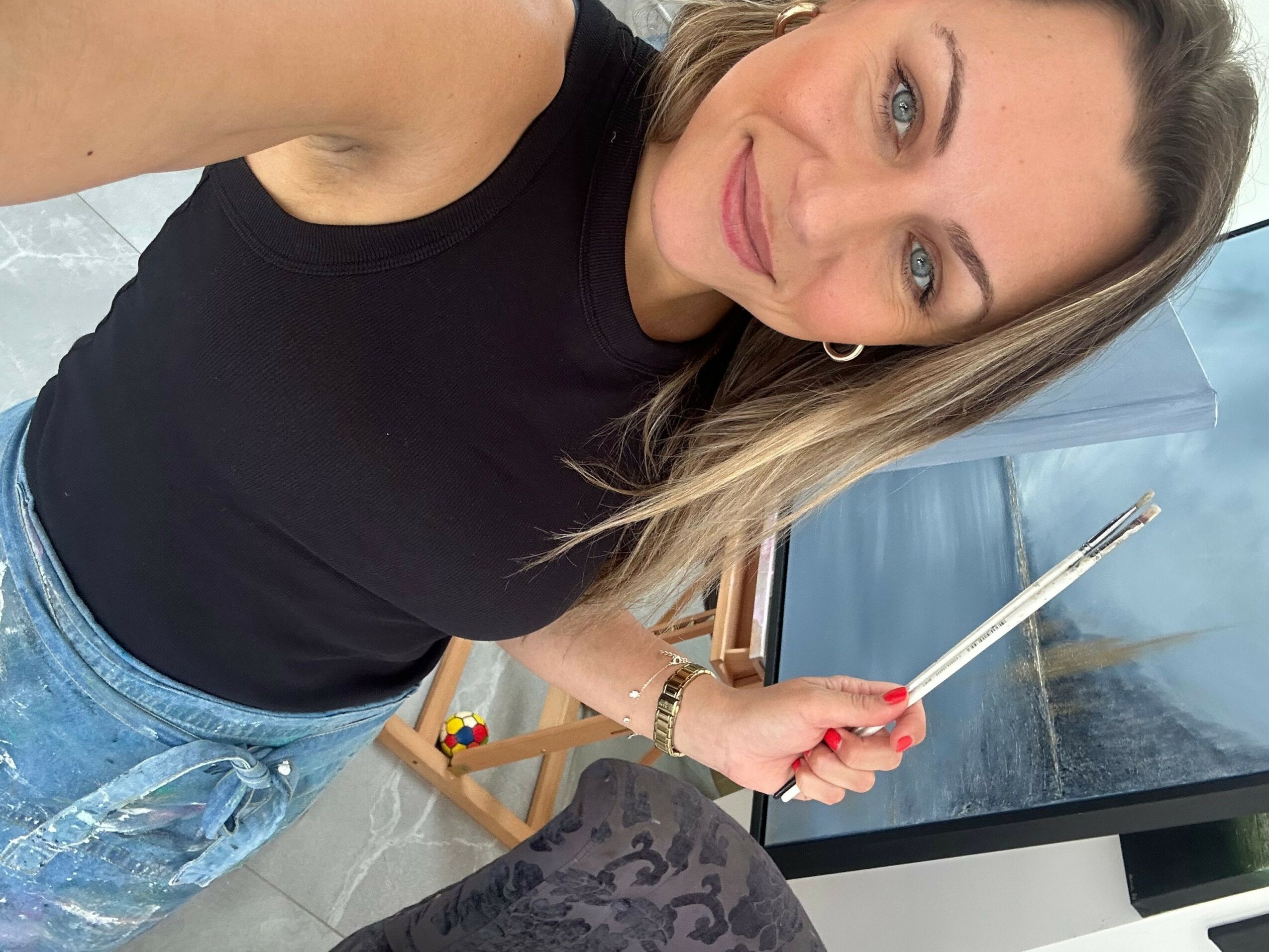 Woman with long blonde hair taking a selfie, wearing a black top and denim shorts, smiling at the camera.