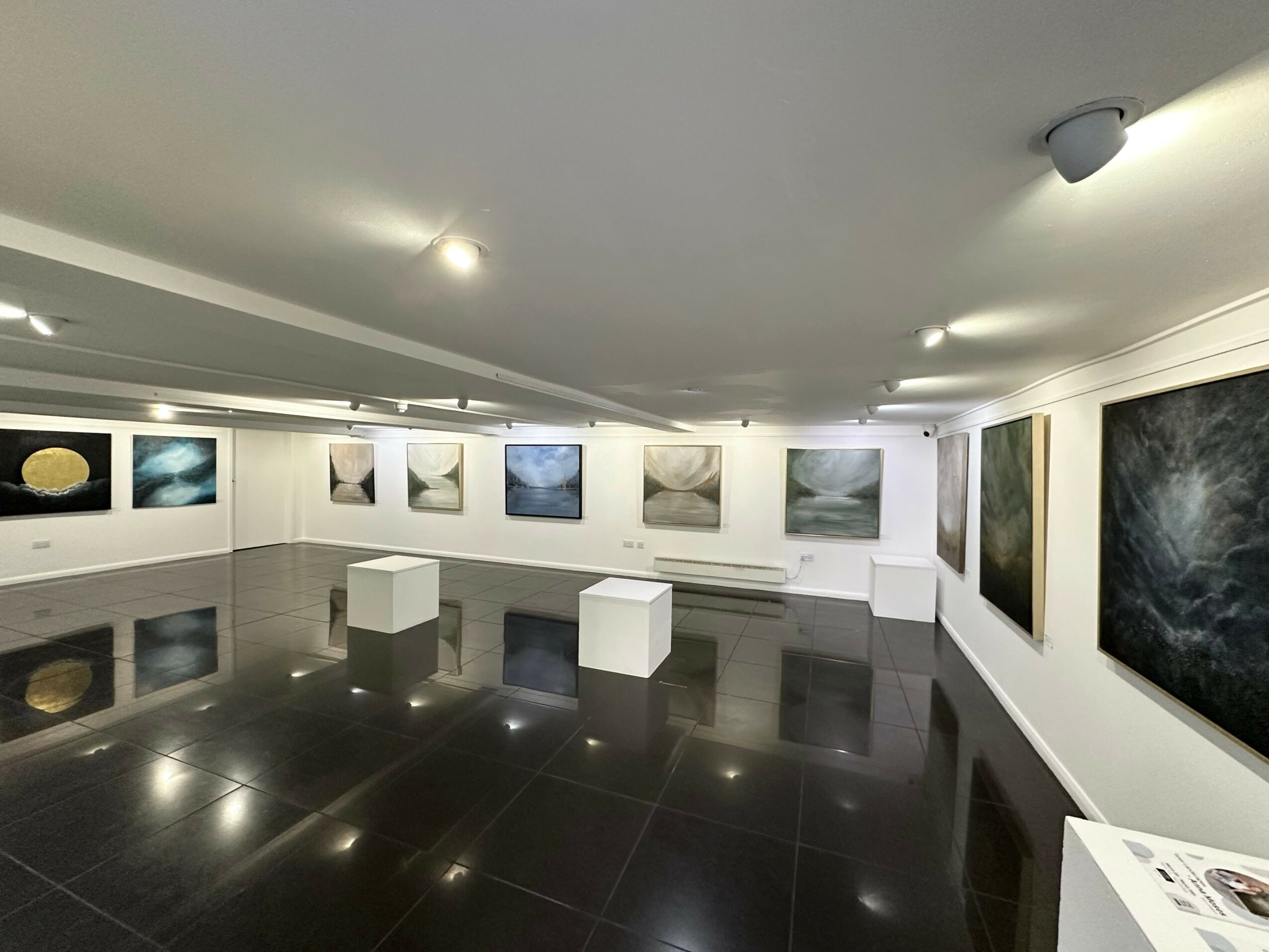 Art gallery with framed paintings on white walls, black tiled floor, and two white benches, ceiling lights illuminating artwork.