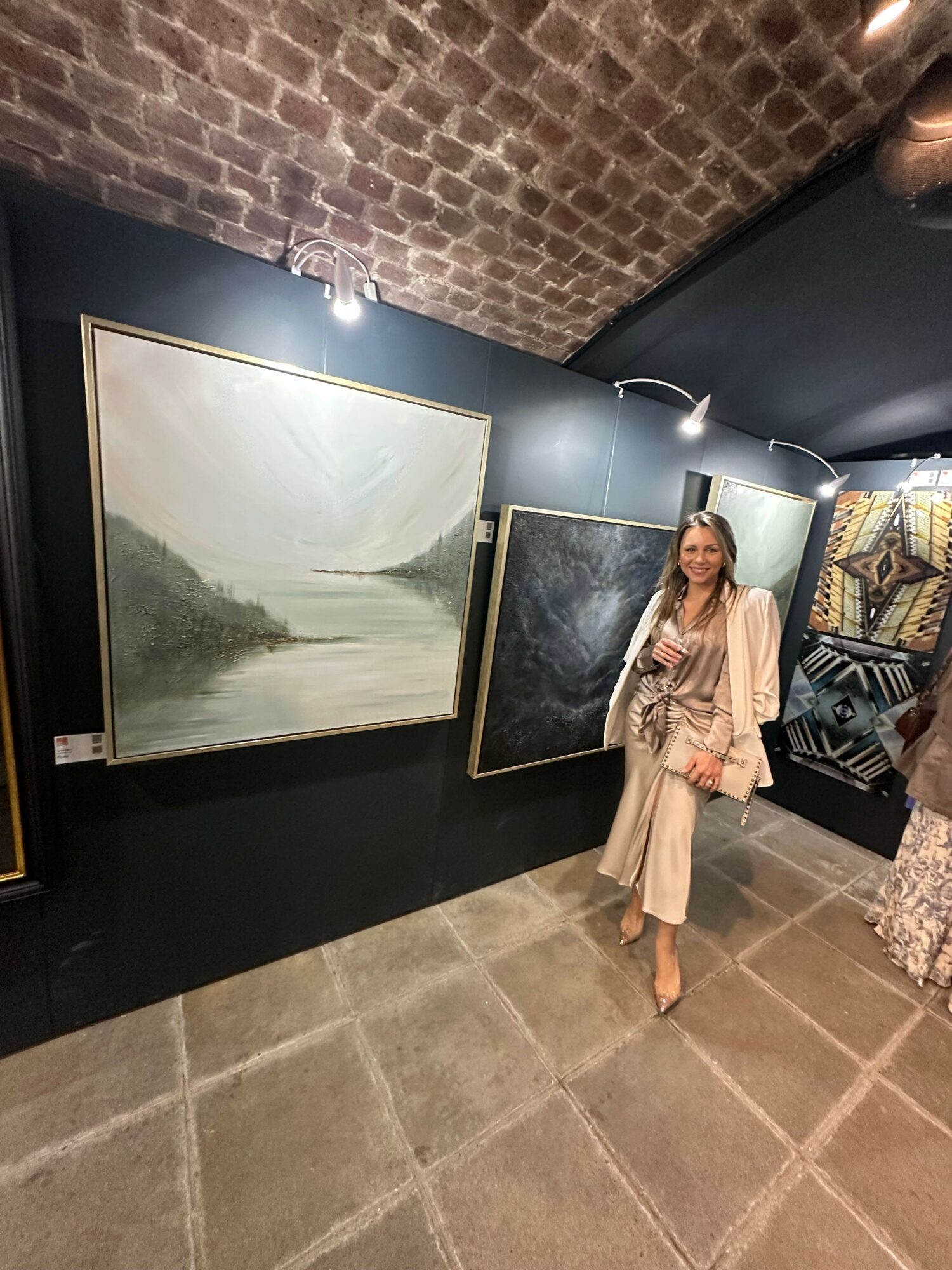 Person standing in an art gallery with paintings on dark walls and tiled floor.