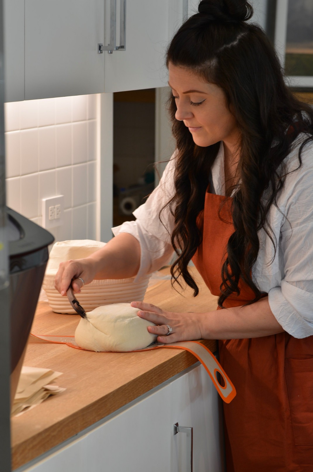 Woman in an apron preparing dough on a kitchen counter, with cabinets and a window in the background.