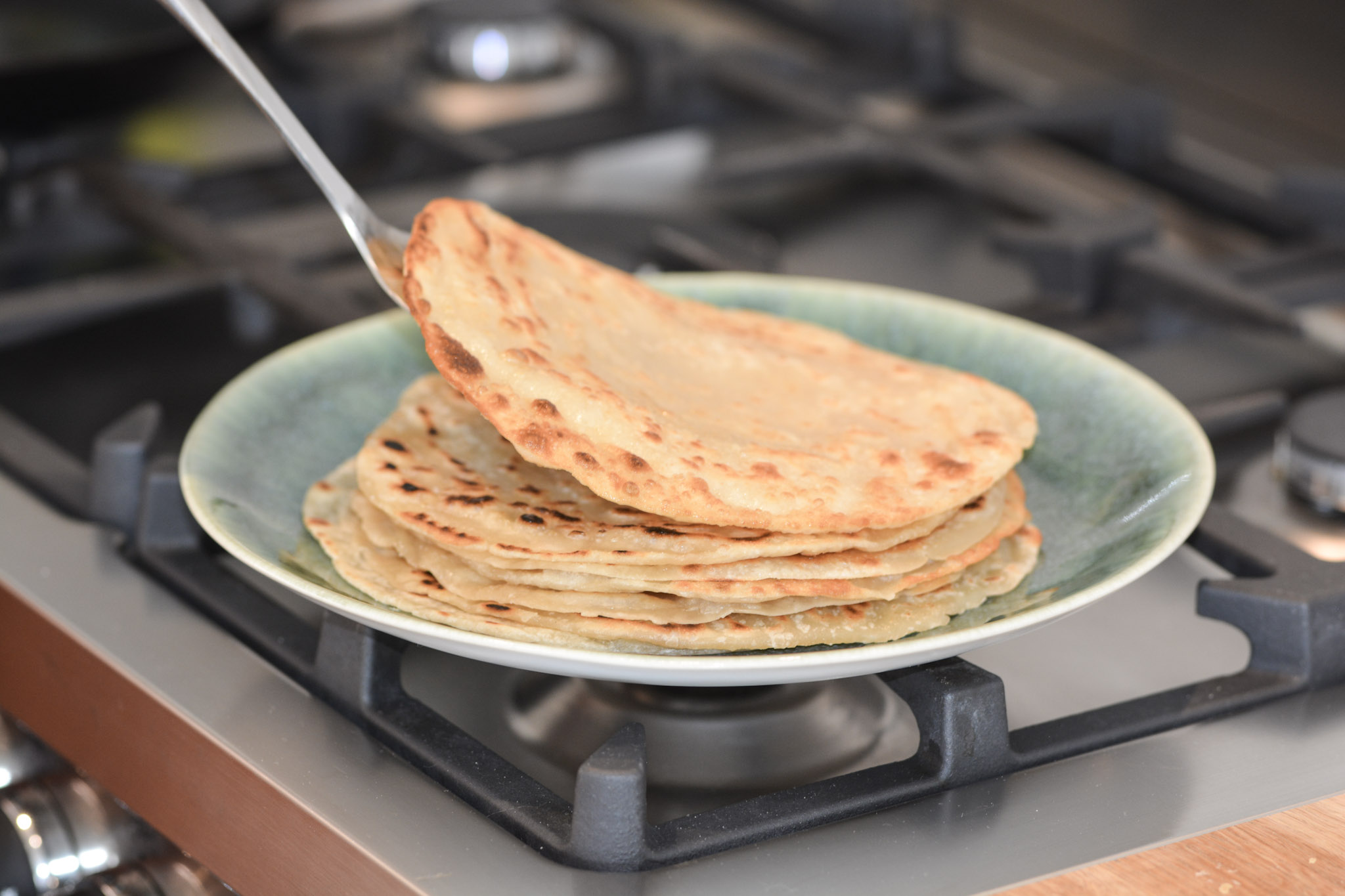 Stack of flatbreads on a green plate over a stove, with one flatbread being lifted.