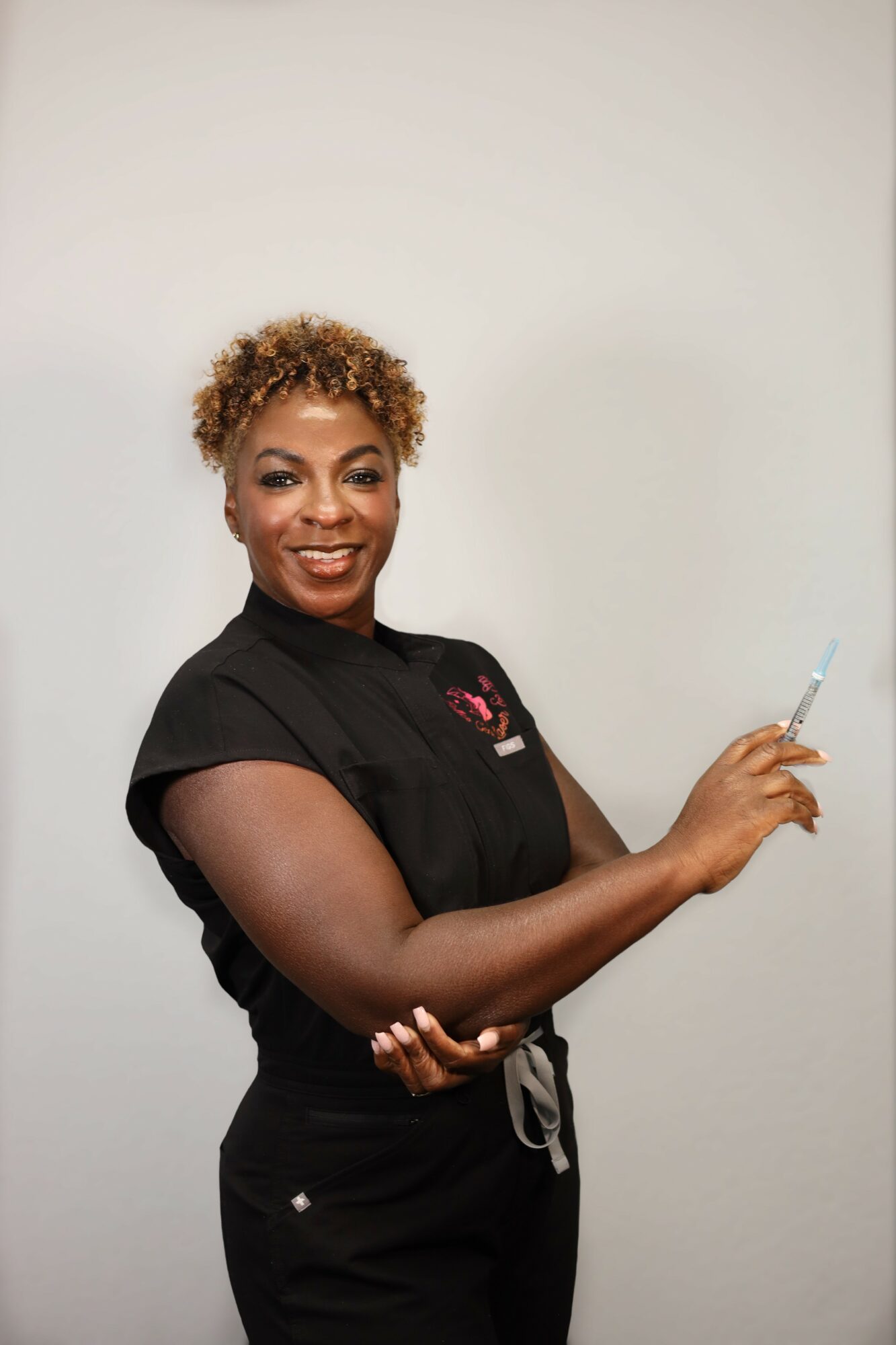 Smiling woman with short curly hair holding a pen, wearing a black medical uniform, standing against a plain background.