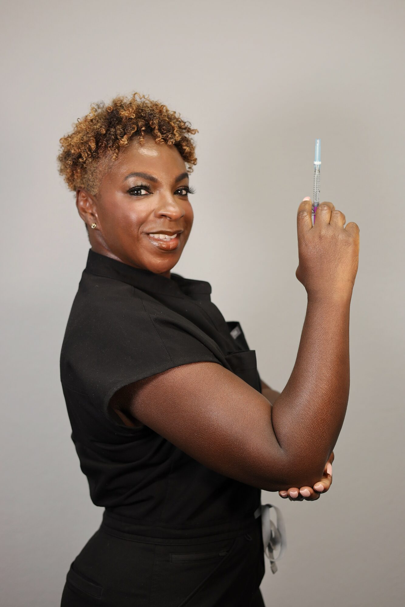 A woman with short curly hair holding a syringe in her right hand, smiling, against a plain background.
