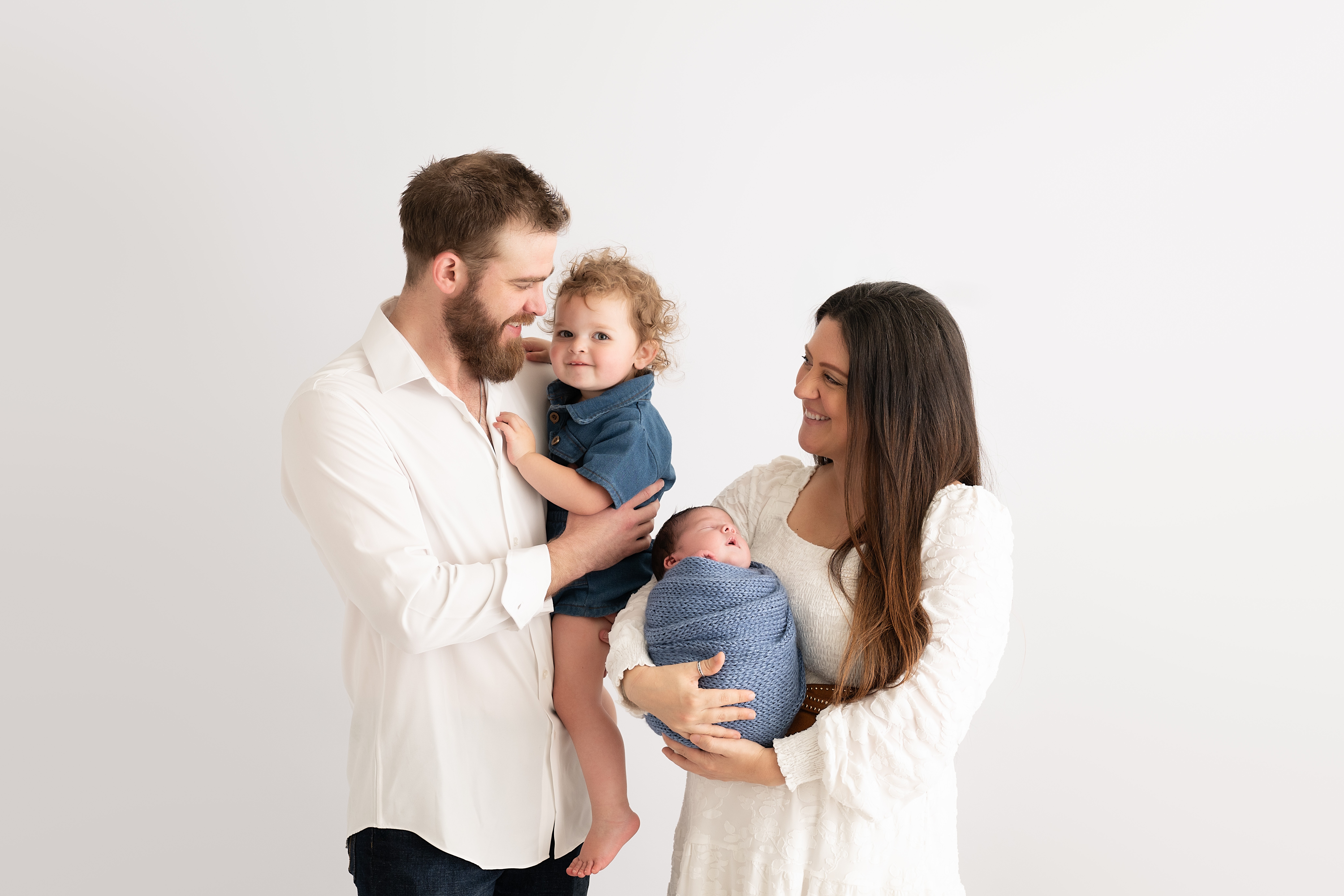 Family of four with a man, woman, young child, and baby, smiling and interacting indoors against a plain background.