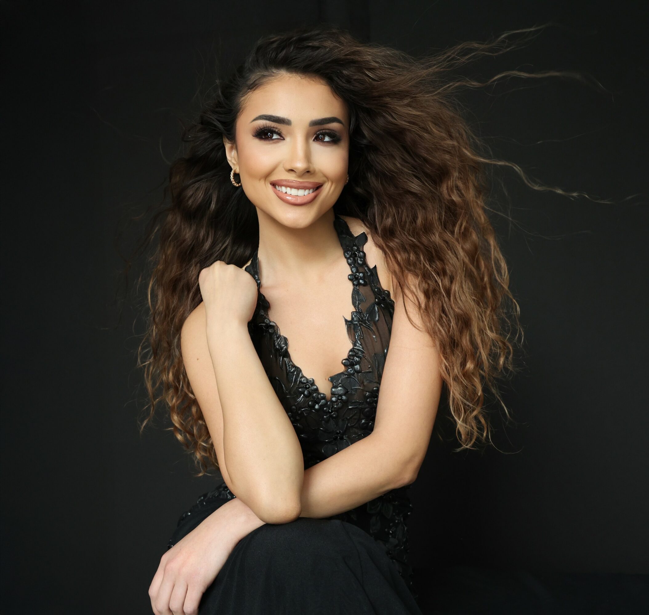 Smiling woman with long, curly hair wearing a black lace top, posing against a dark background.