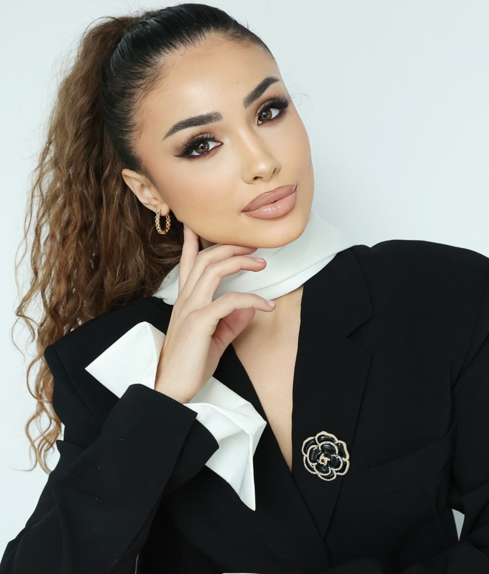 Woman with makeup and curly hair, wearing a black blazer with a white scarf, posing with her hand near her face.