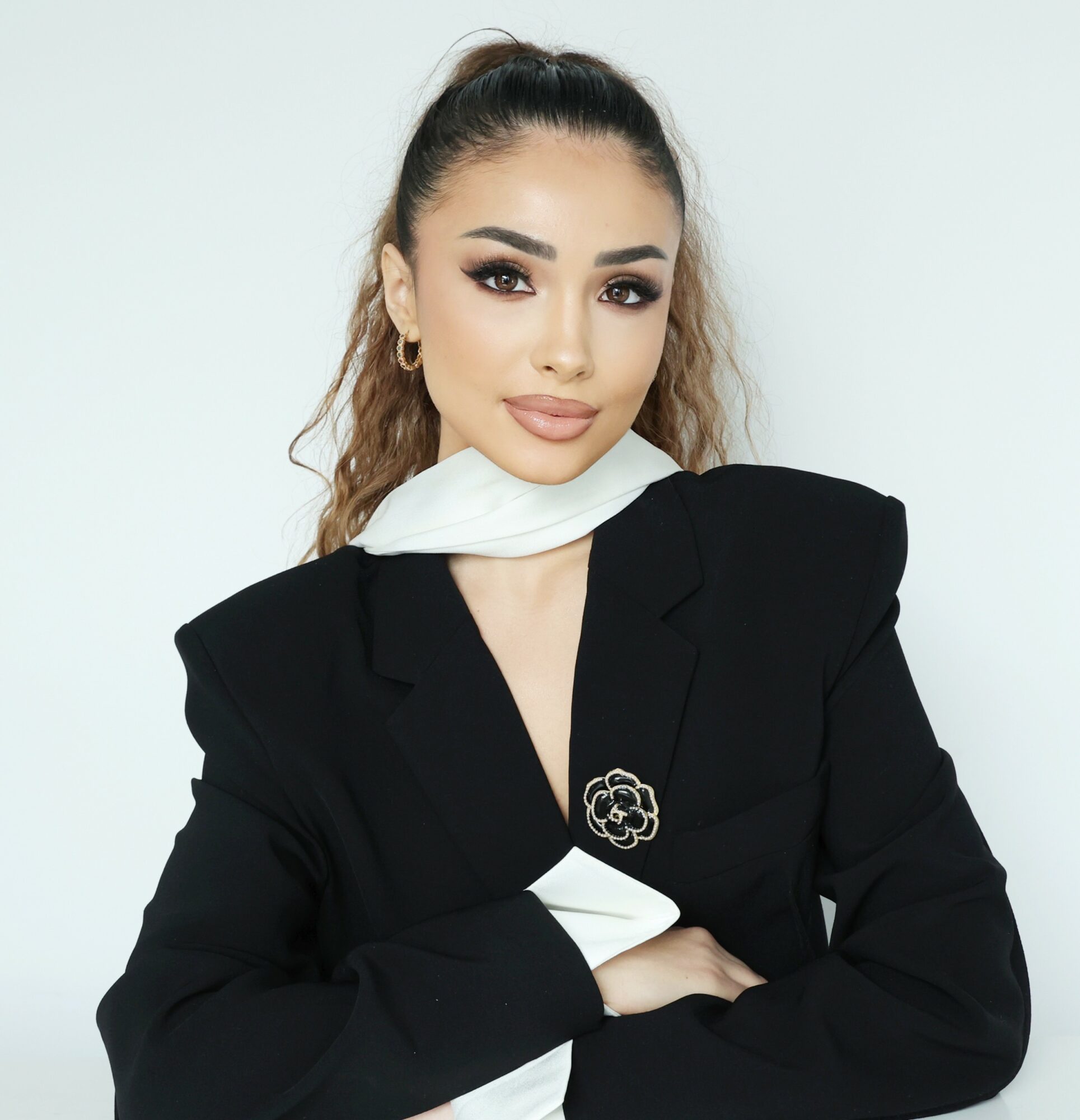 Woman with dark hair in a high ponytail, wearing a black blazer and white scarf, looking at camera.