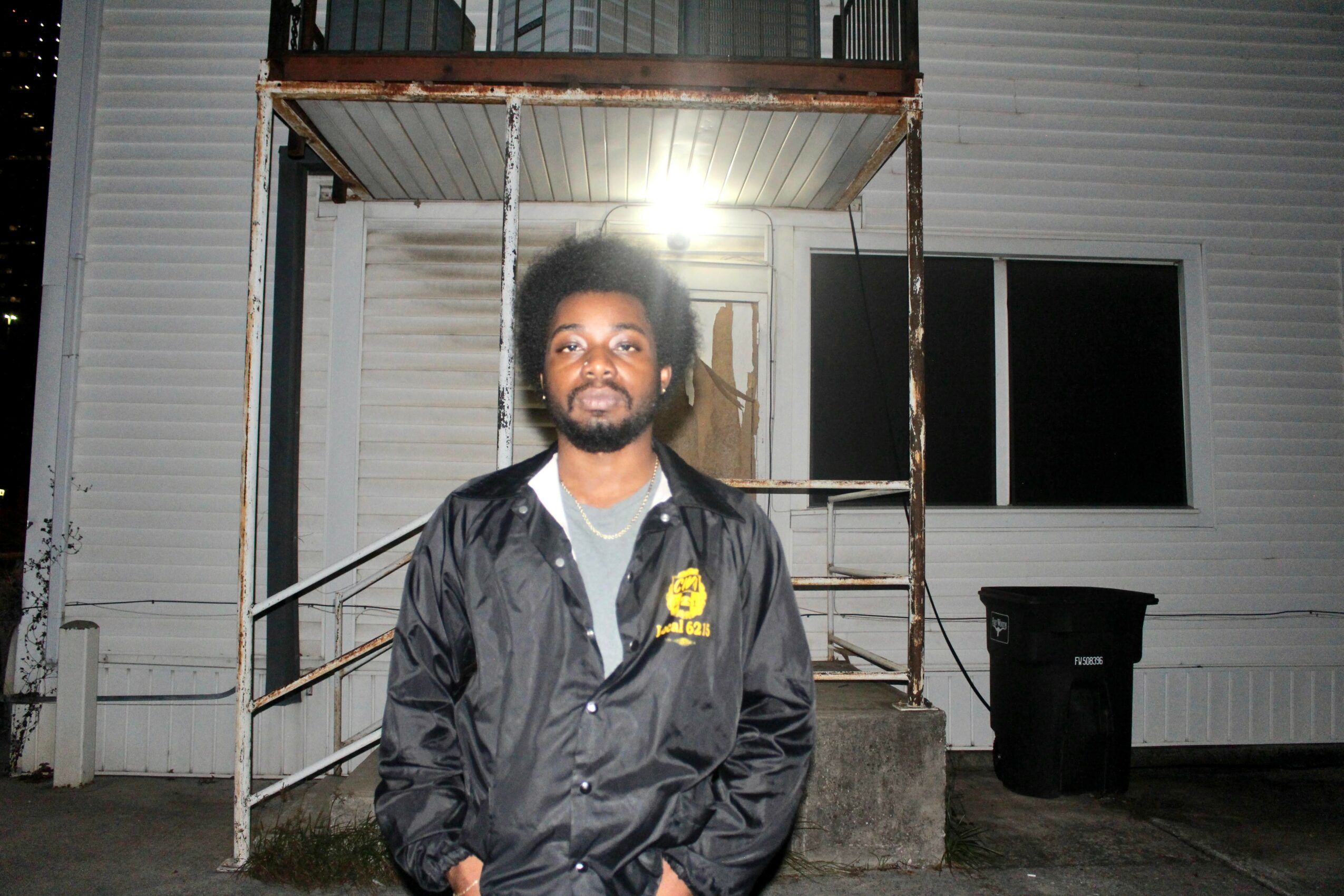 Young man with curly hair and beard standing outside a building at night, wearing a black jacket with a yellow logo.