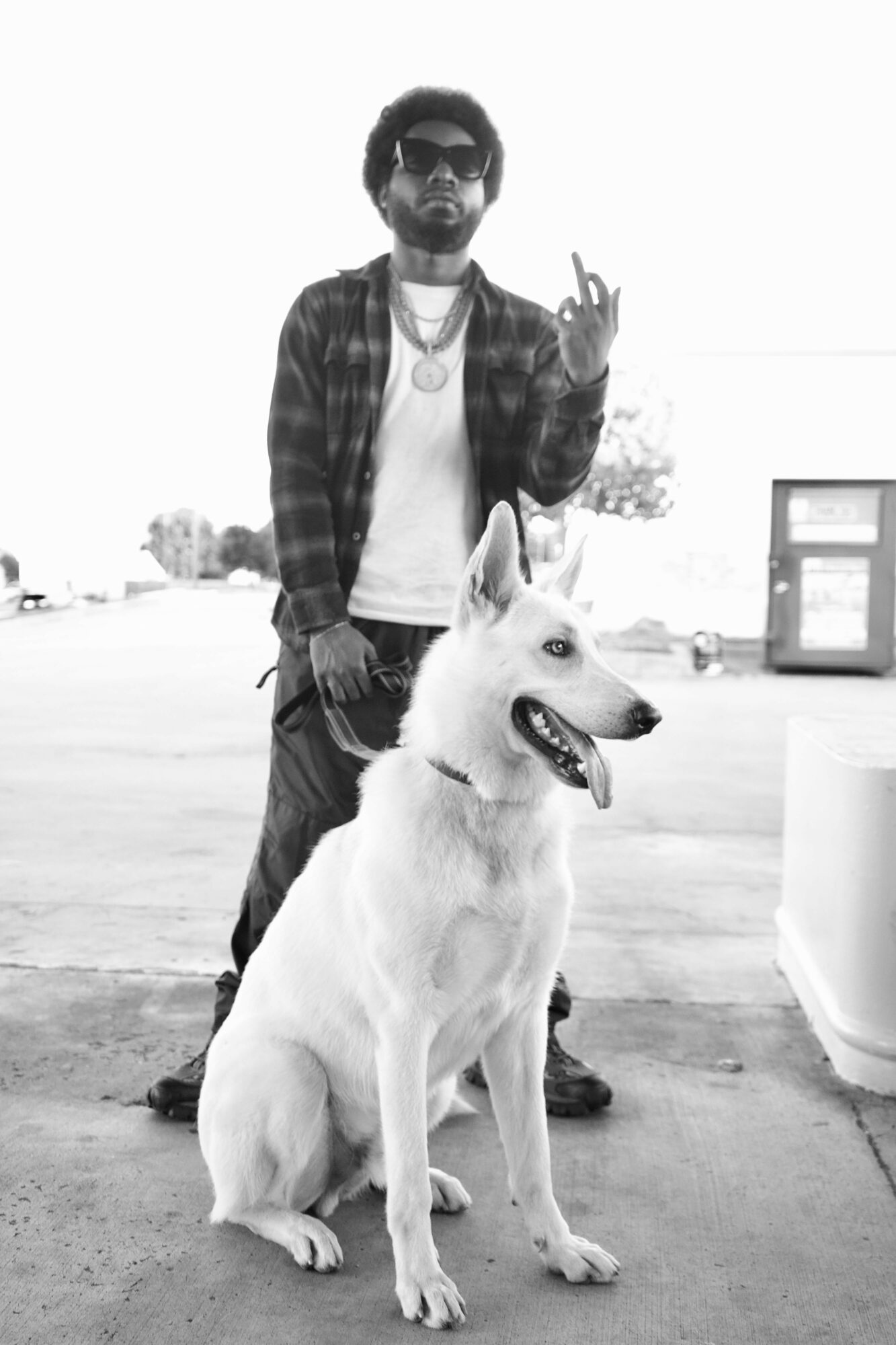 Man with sunglasses and a dog outdoors, making a gesture with his hand, in black and white.