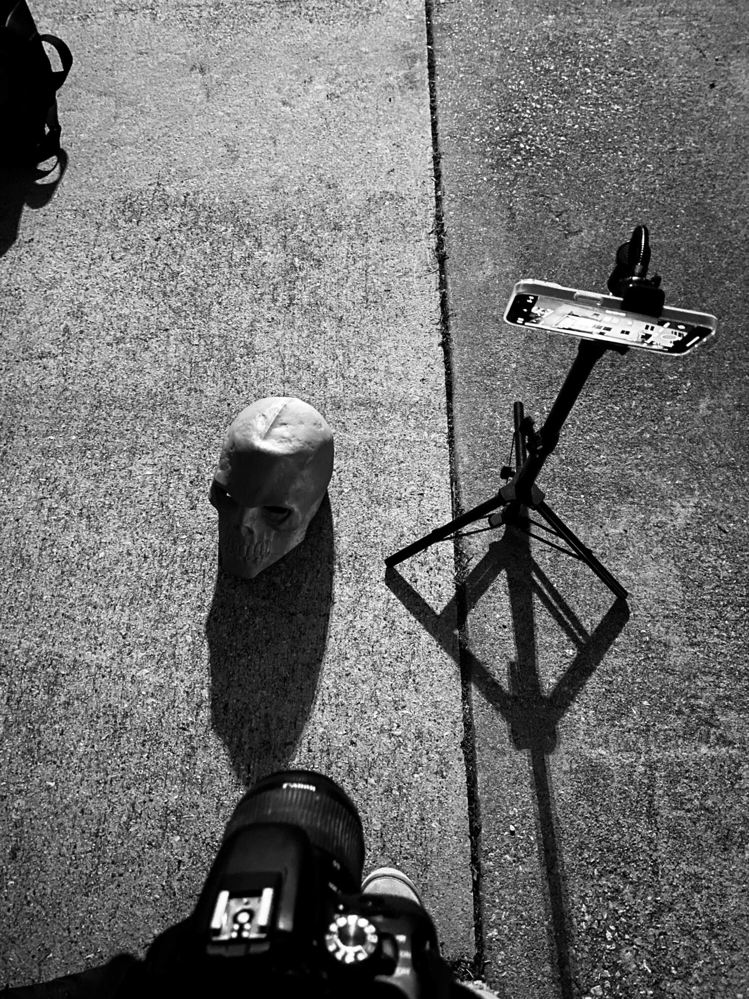 A smartphone on a tripod captures a scene with a small object and a camera on a concrete surface.