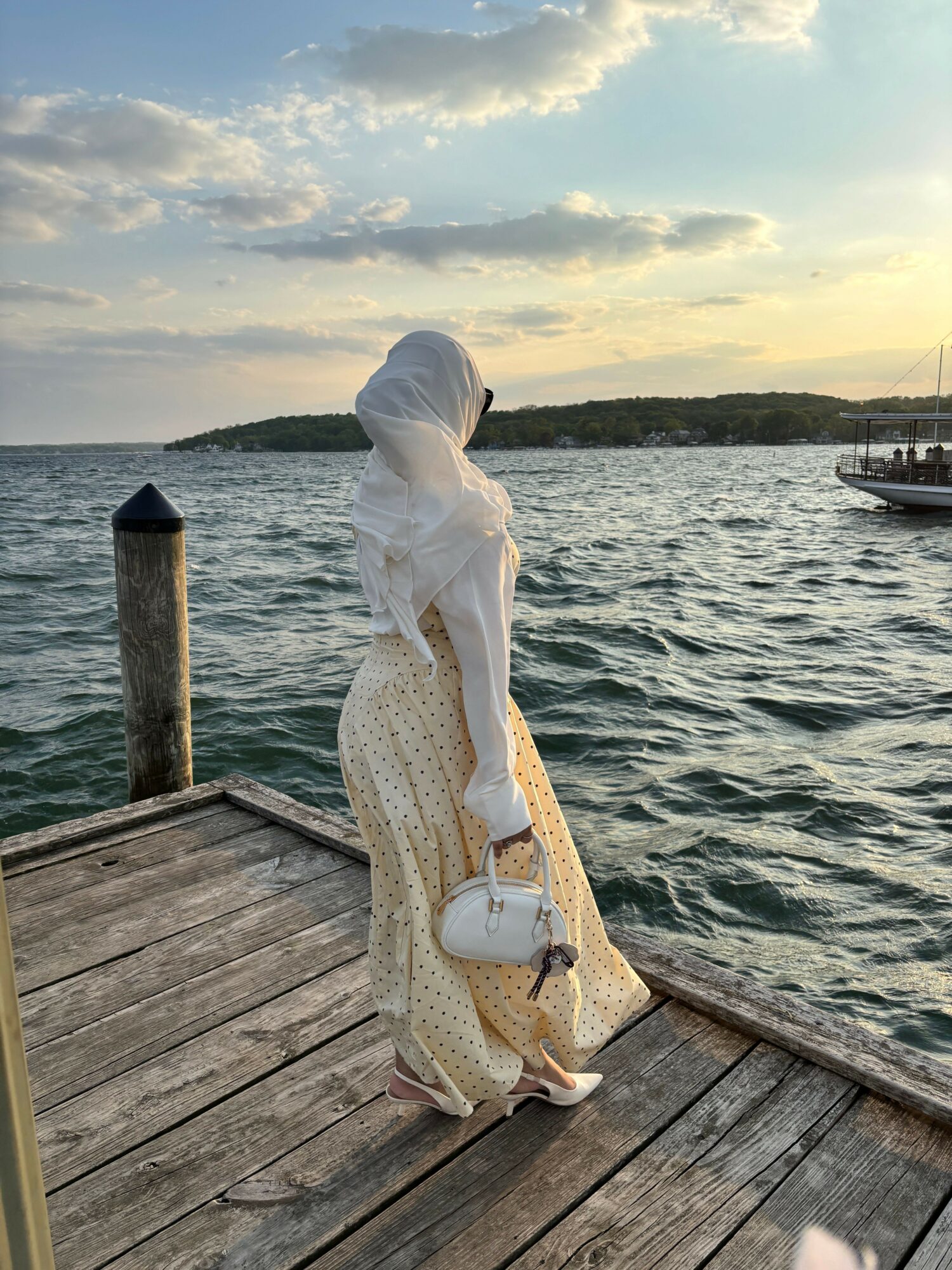 Woman in white dress and headscarf standing on wooden dock by water, holding a glass of wine, with cloudy sky and boats in background.