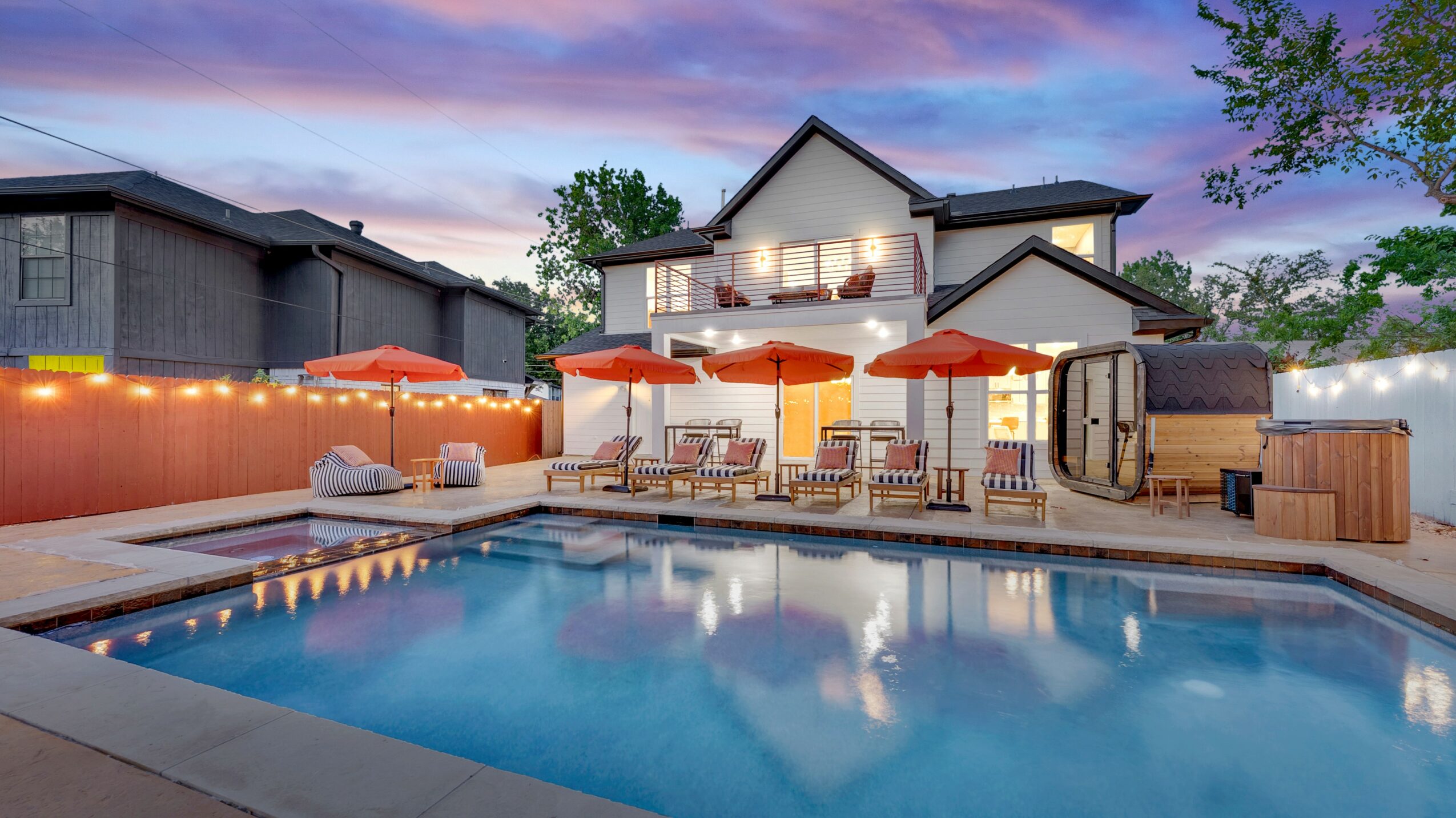 Backyard with swimming pool, patio area with tables and umbrellas, and a two-story house in the background at sunset.
