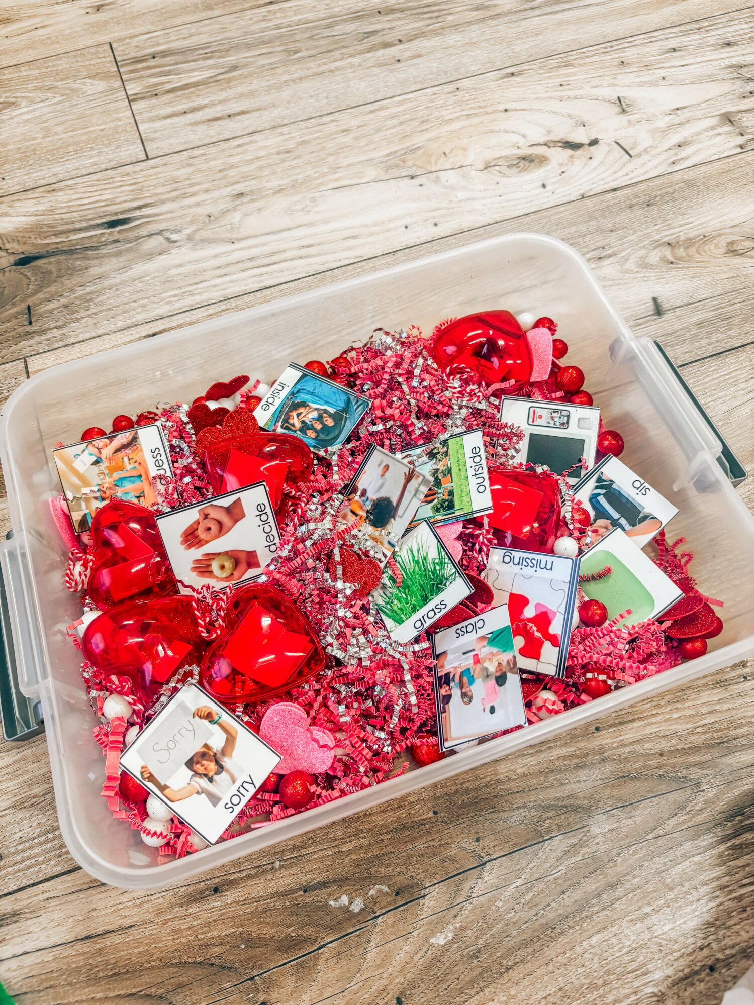 Clear plastic container filled with red and pink Christmas ornaments and small gift cards on a wooden surface.
