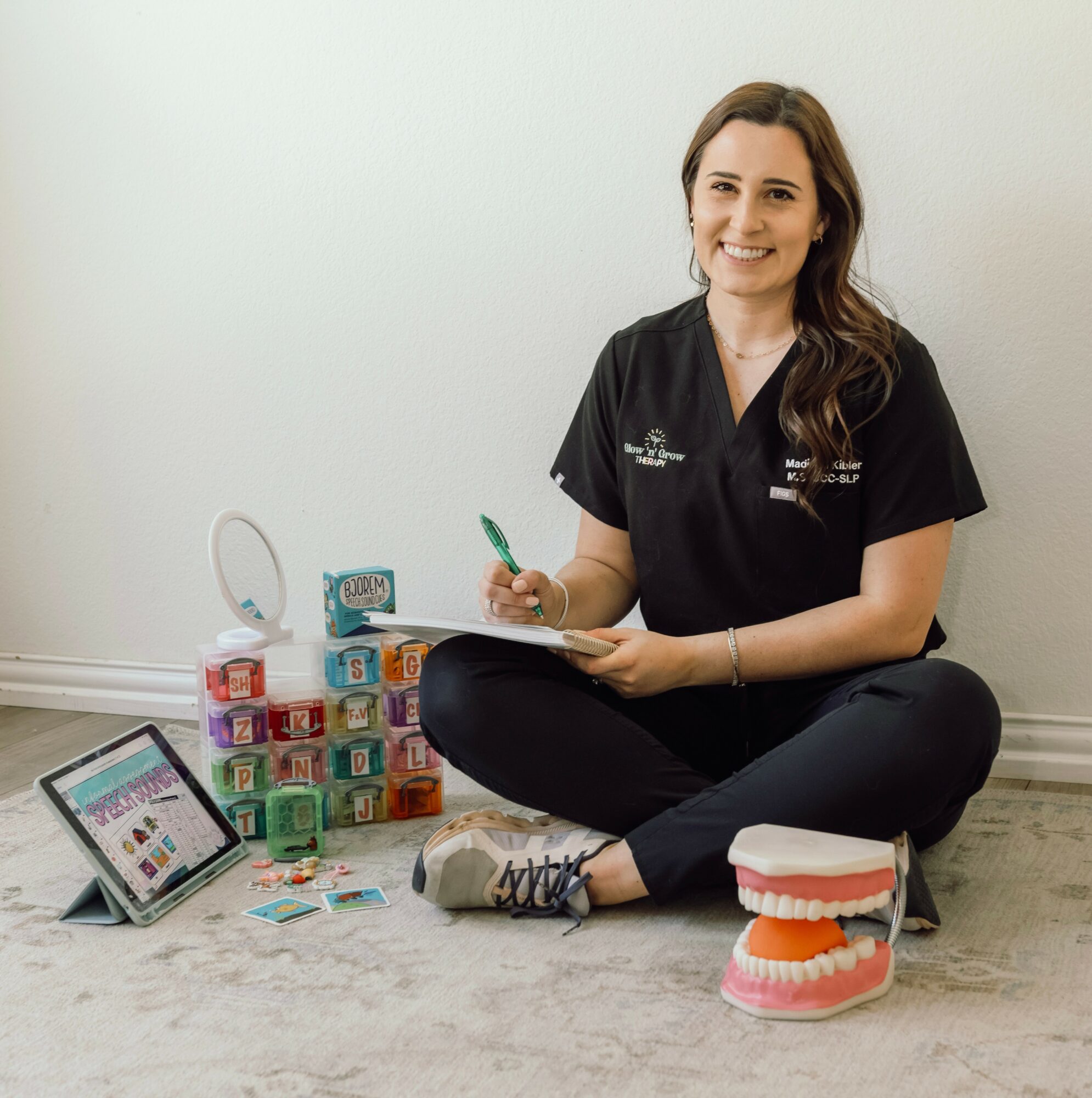 Woman sitting on floor with dental models, tablet, and colorful blocks, smiling and holding a green pen, against a plain wall.