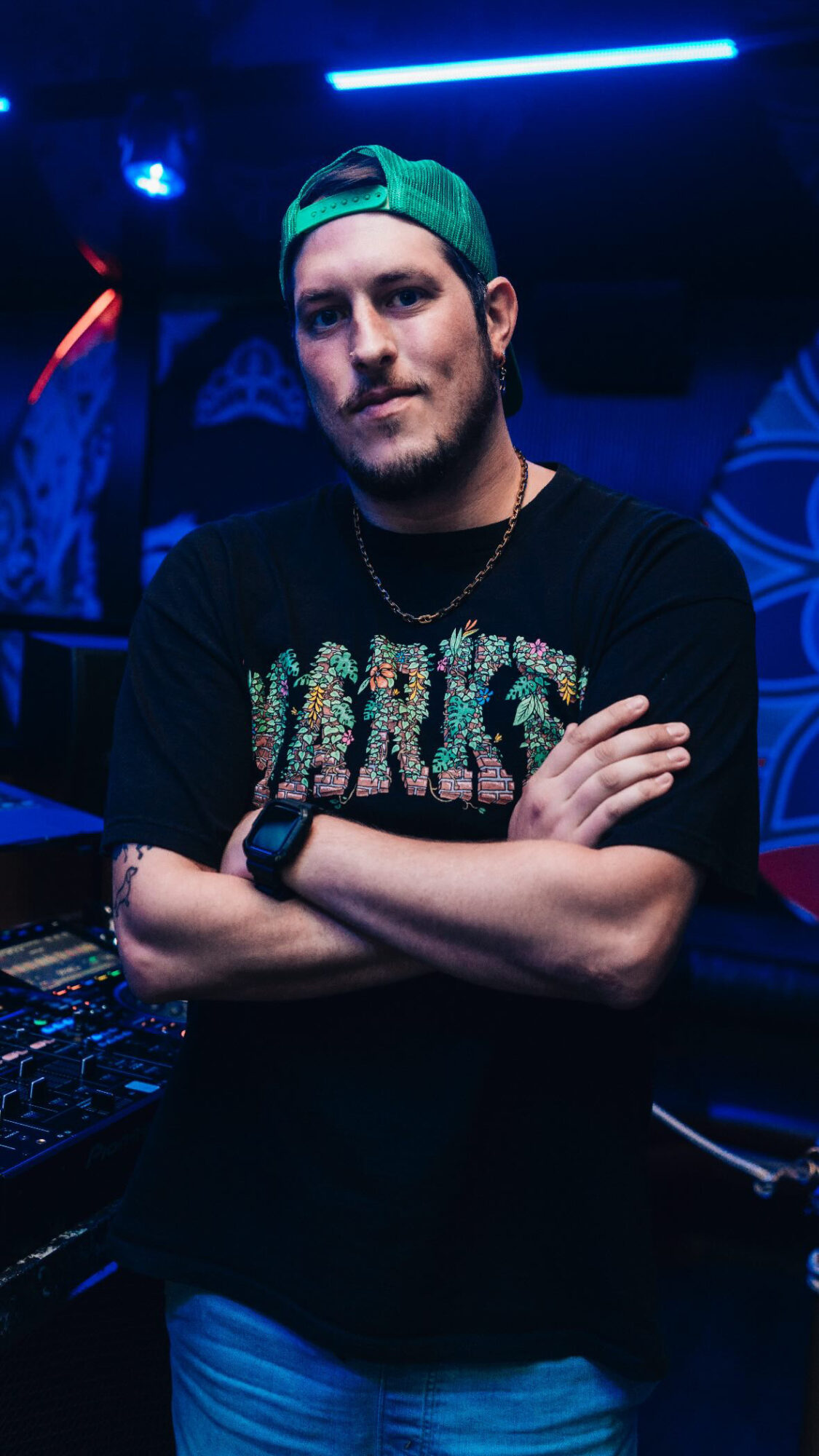 Young man with a beard wearing a green cap backwards, black shirt, and a watch, standing with arms crossed in a club.