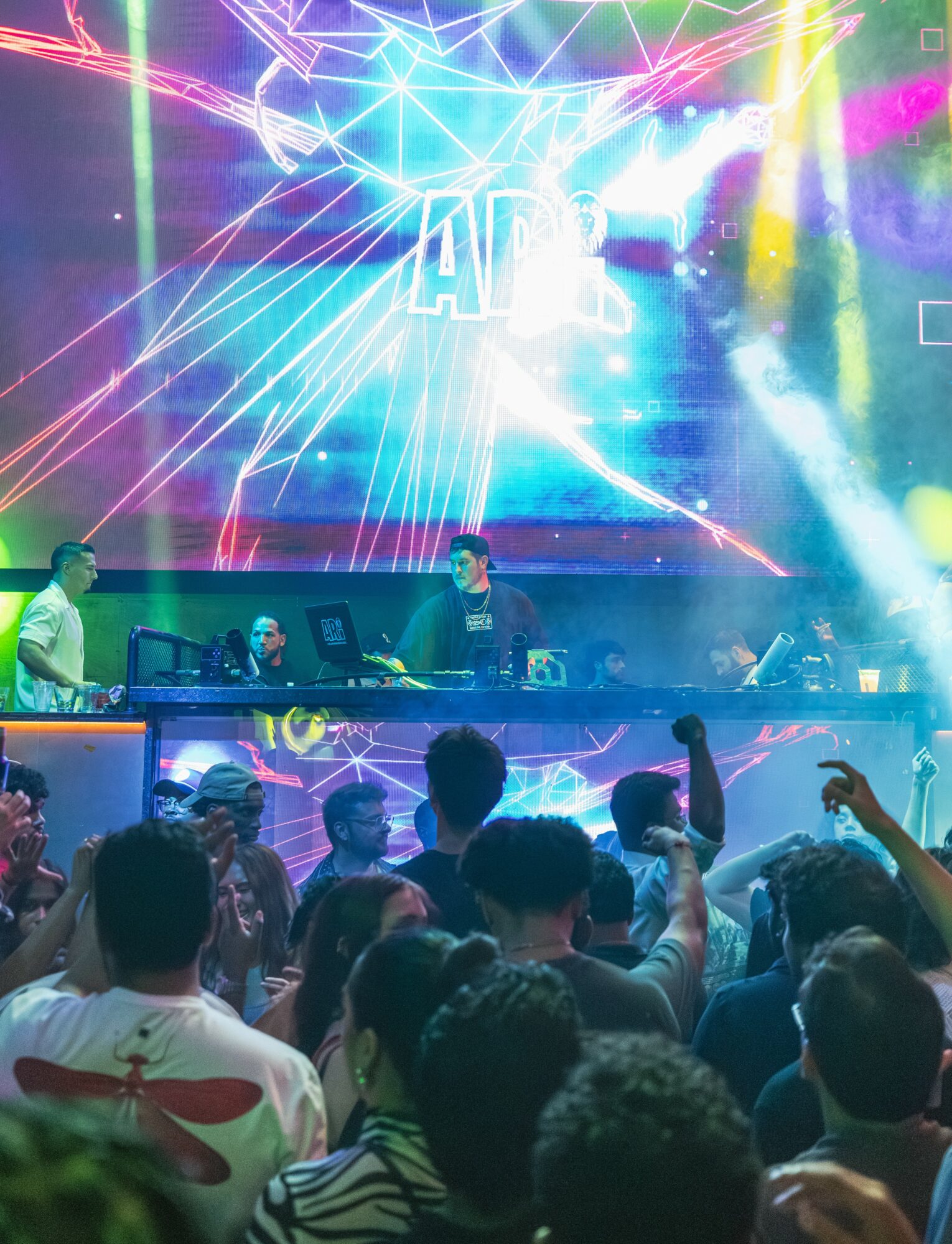 Crowd dancing and DJ performing on stage with colorful lights and visual effects in the background.