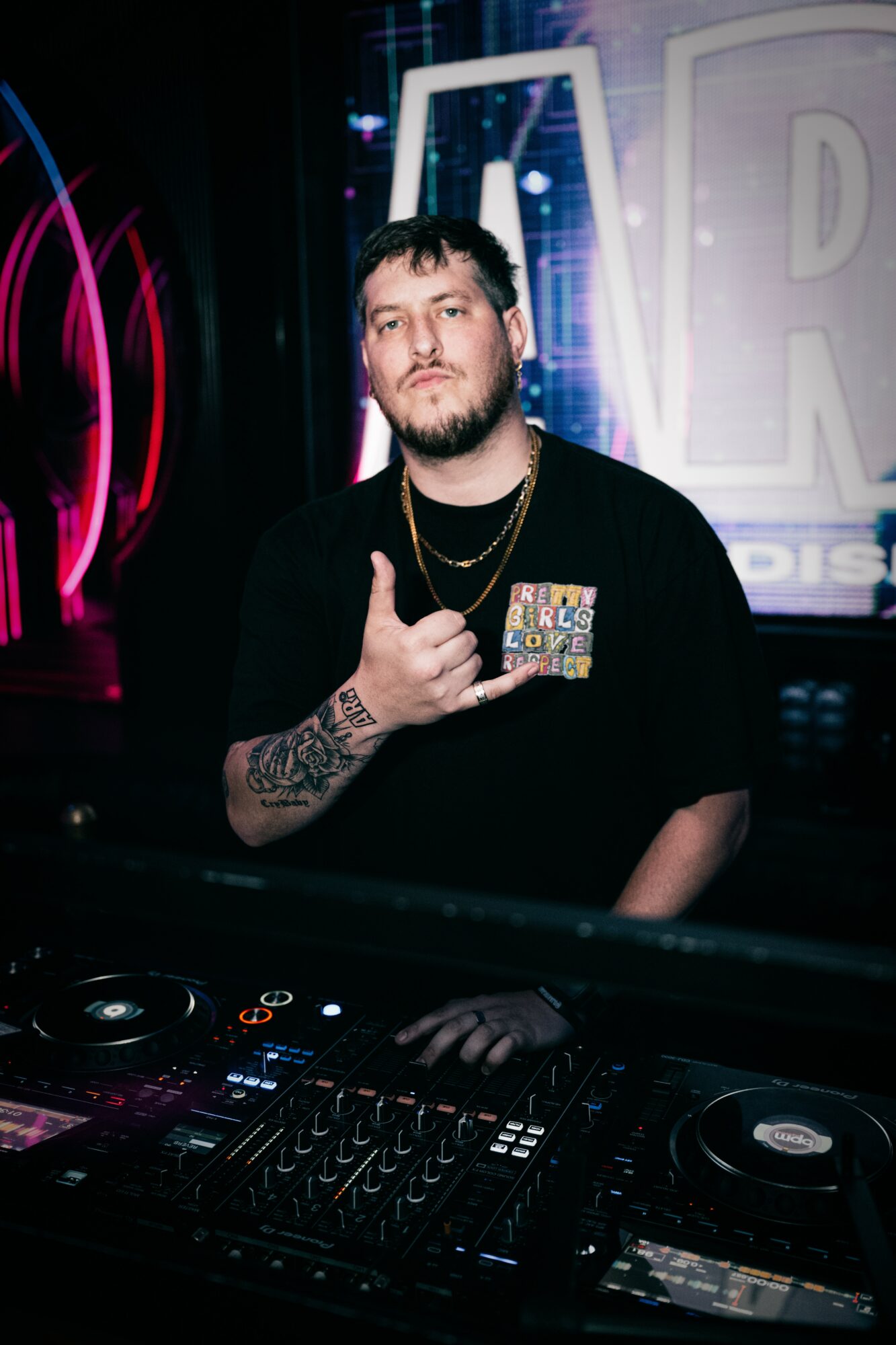 Person with dark hair and beard, wearing black shirt and gold chains, standing behind DJ equipment in a dark club setting