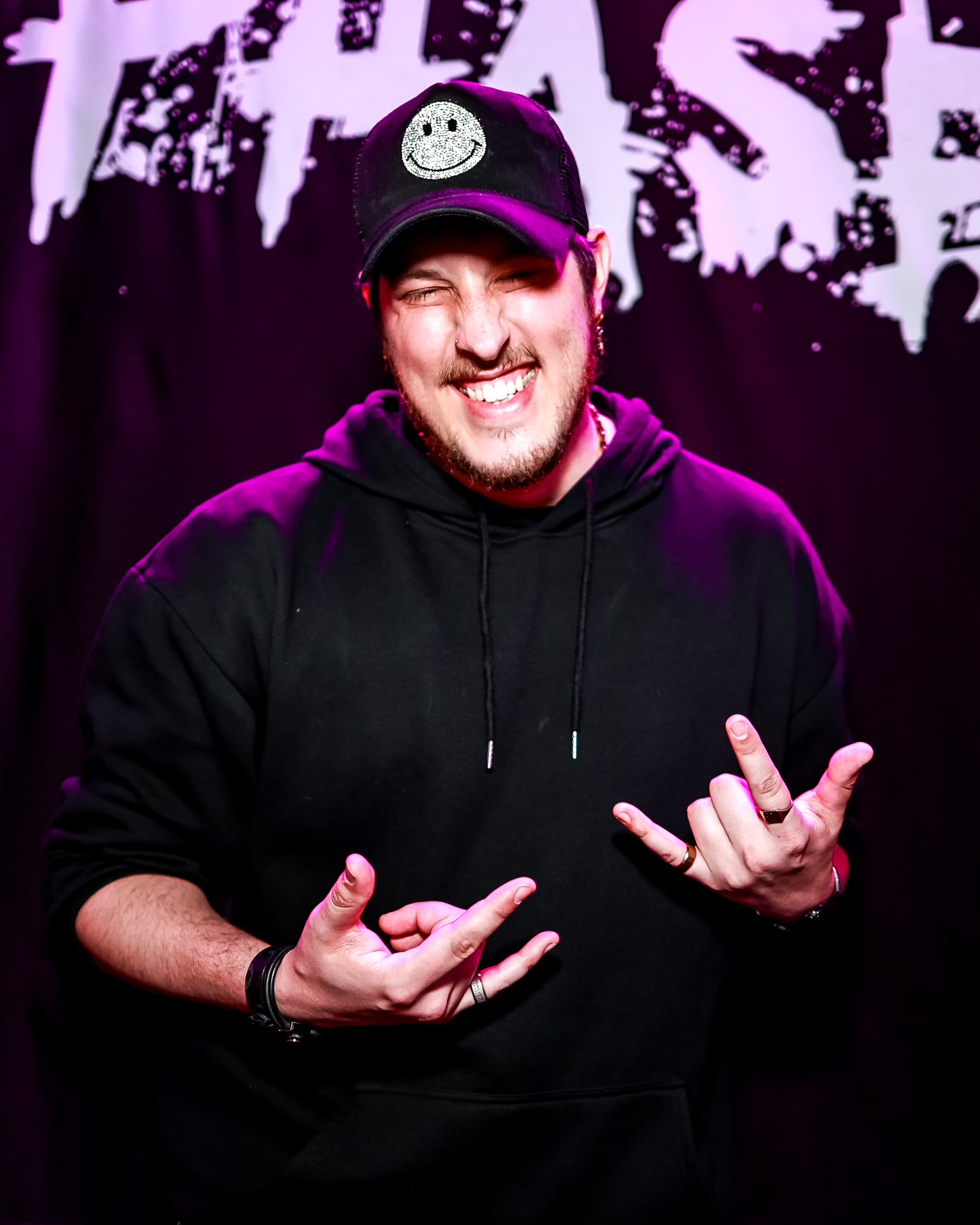 Young man smiling, wearing a black hoodie and cap, making hand gestures, dark background with white text
