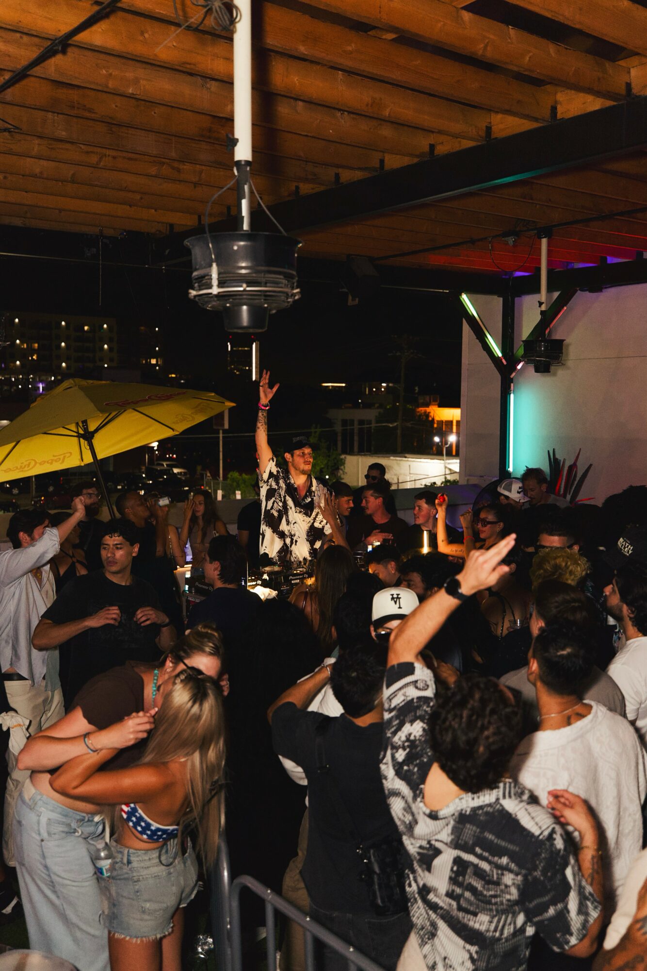 Crowd of people at a nighttime outdoor event, some raising hands, with a person on stage under a ceiling with wooden beams.