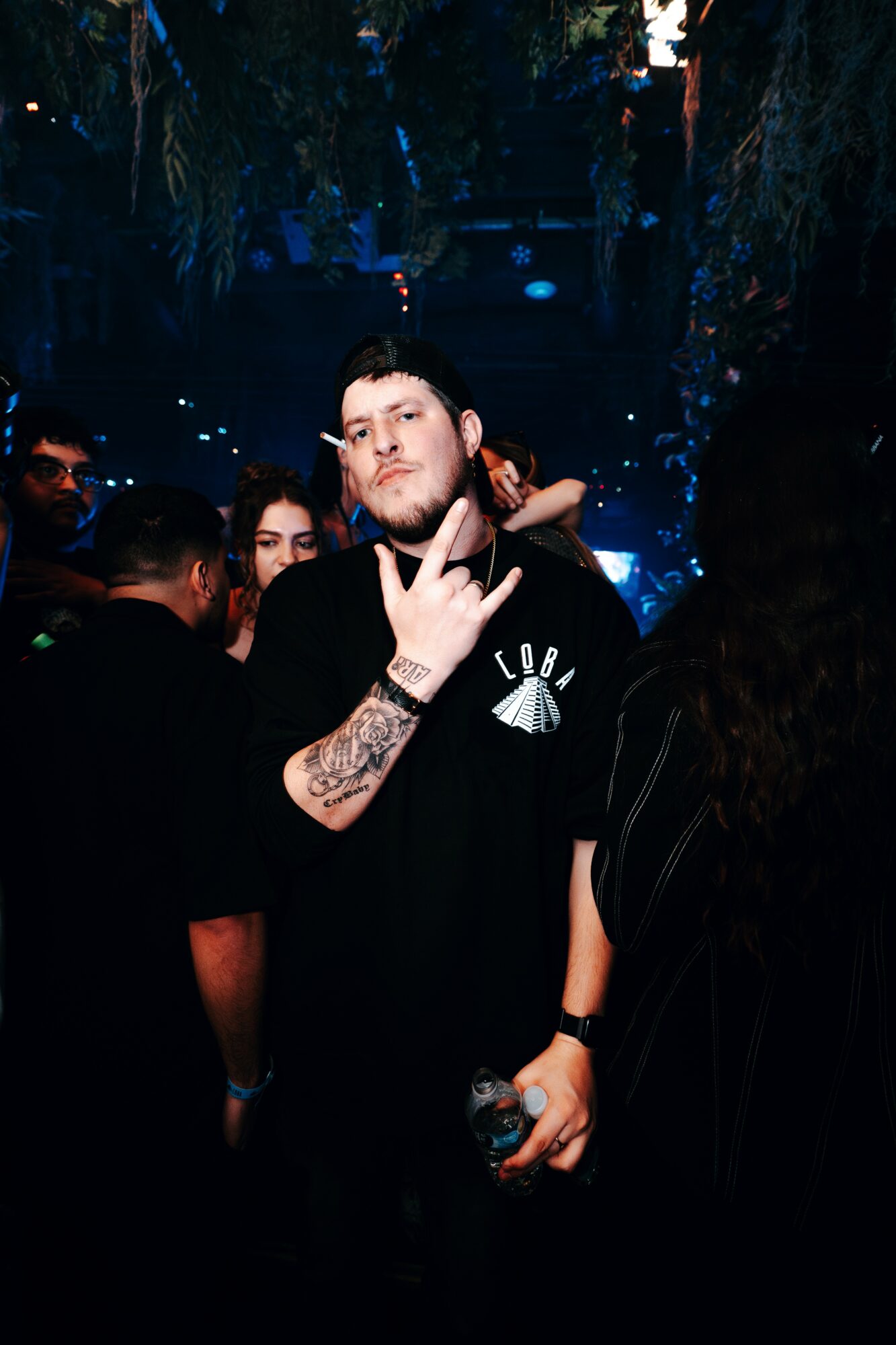 Young man with tattoos making a hand gesture in a dark, crowded party setting with blue lighting.