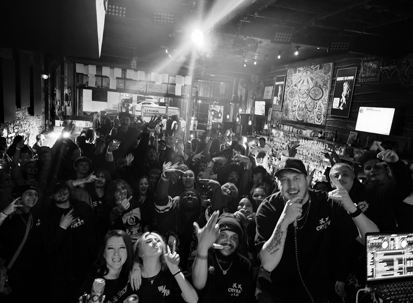 Crowd of people in a bar or club with a DJ, some smiling and raising hands, illuminated by bright lights.