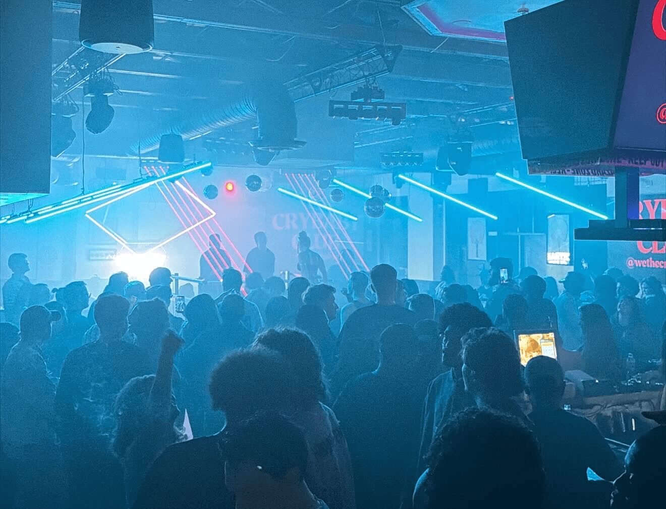 Crowd of people in a nightclub with colorful laser lights and a DJ stage, some holding phones, dark atmosphere.