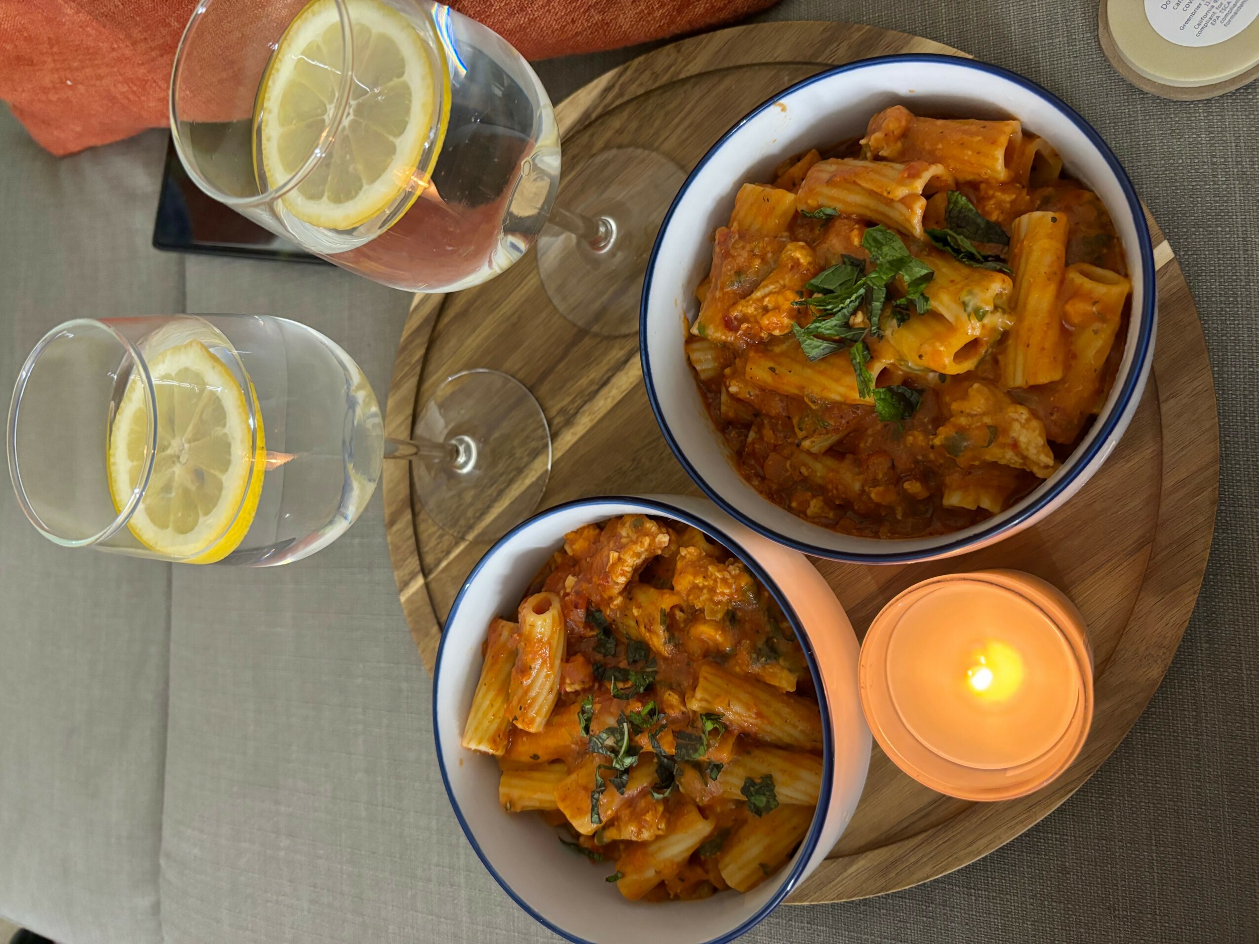 Two bowls of pasta with sauce and herbs on a wooden tray, two glasses of lemon water, and a lit candle.