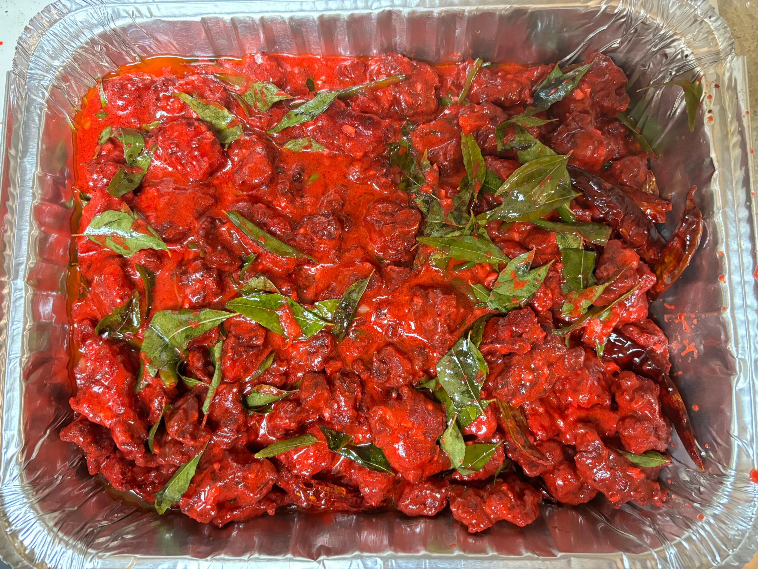 Red cooked meat with green herbs in a foil container, garnished with leafy herbs, close-up view.