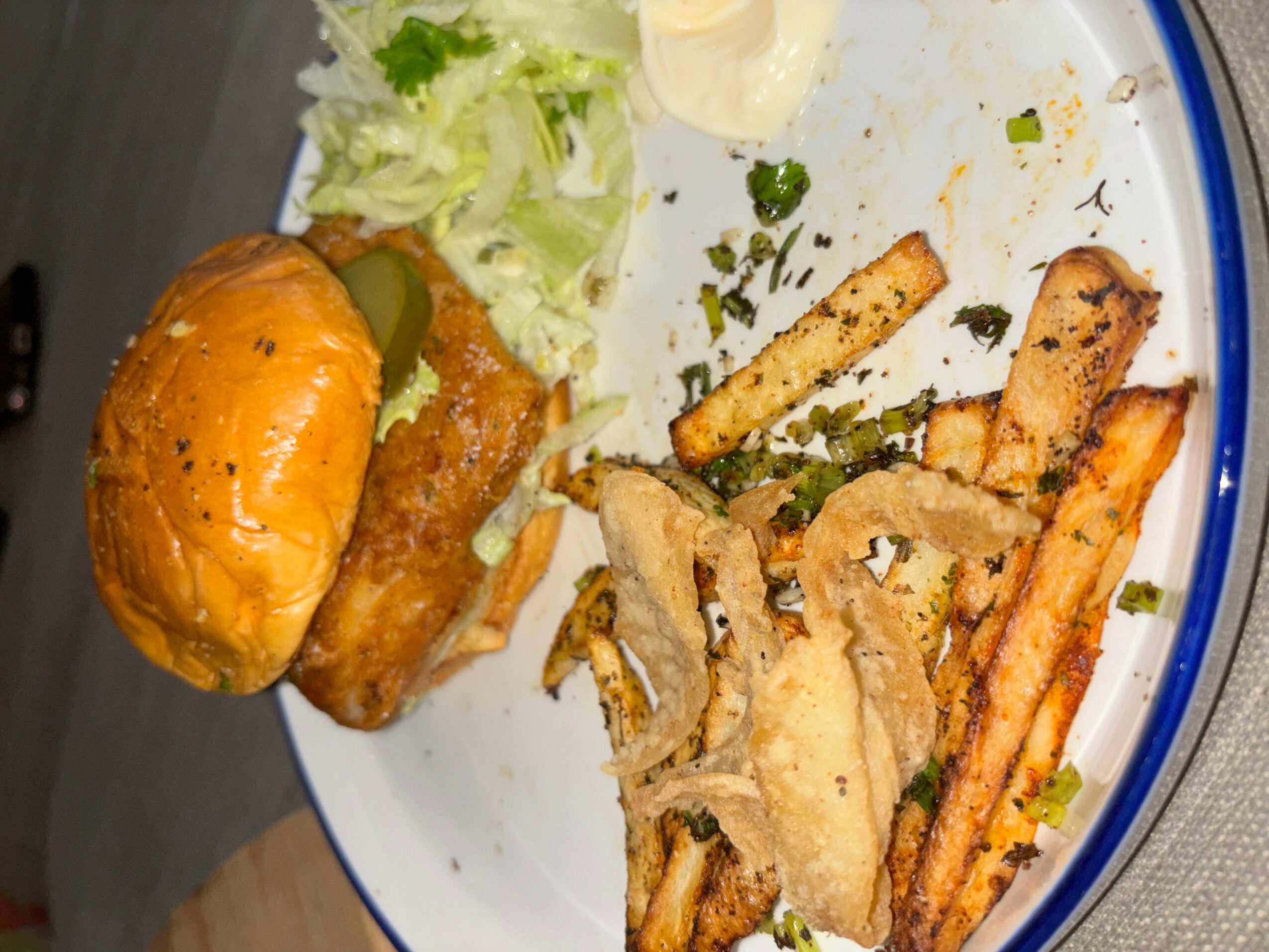 Burger with lettuce, fried fish, and fries on a white plate, with some salad and sauce on the side.