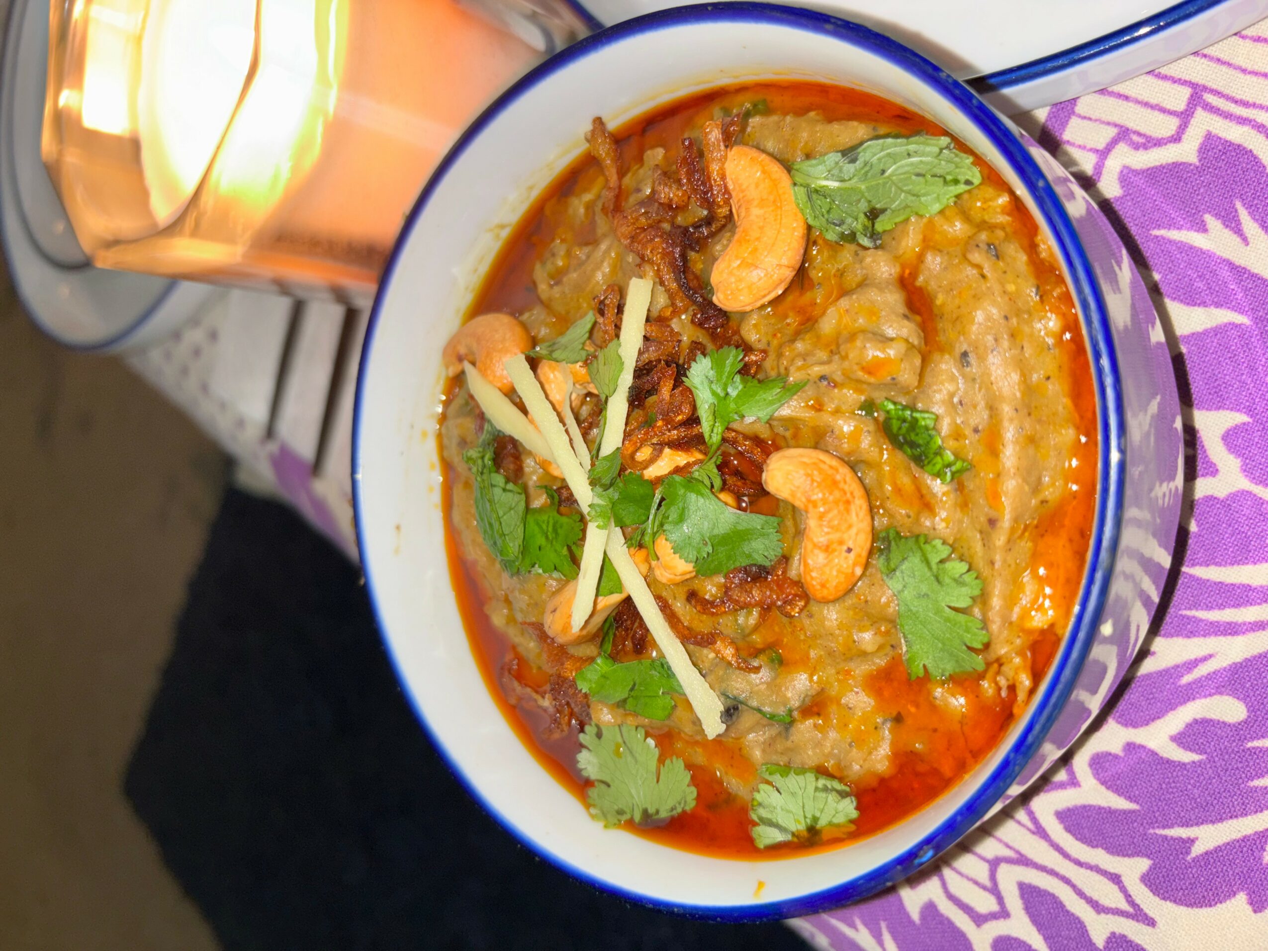 Bowl of spicy curry with shrimp, herbs, and vegetables, viewed from above, on a purple patterned surface.