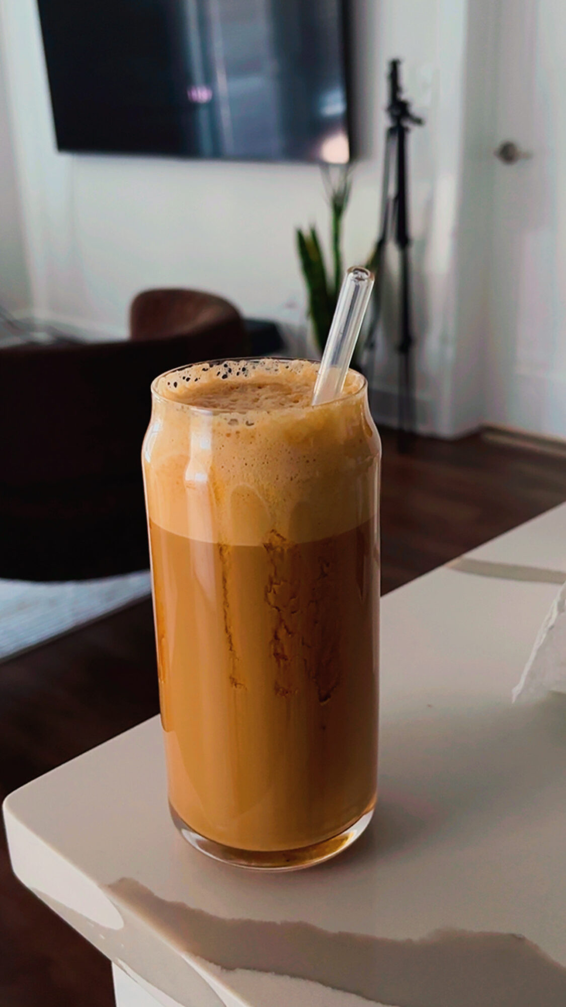 Tall glass of frothy iced coffee with a straw on a white table, background includes a TV and plant.