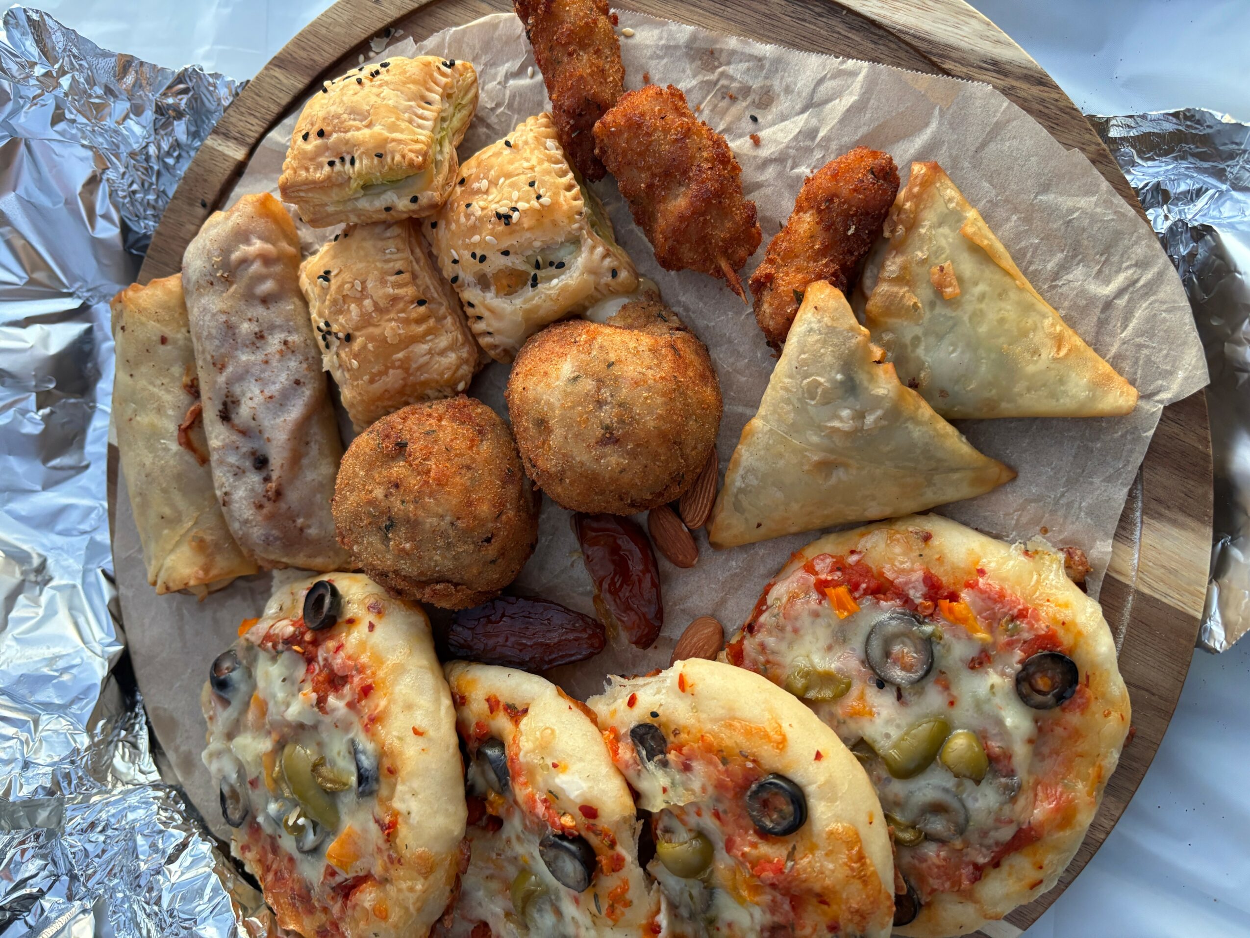 Assorted fried and baked snacks on a wooden platter, including pizza slices, breaded sticks, and fried balls.