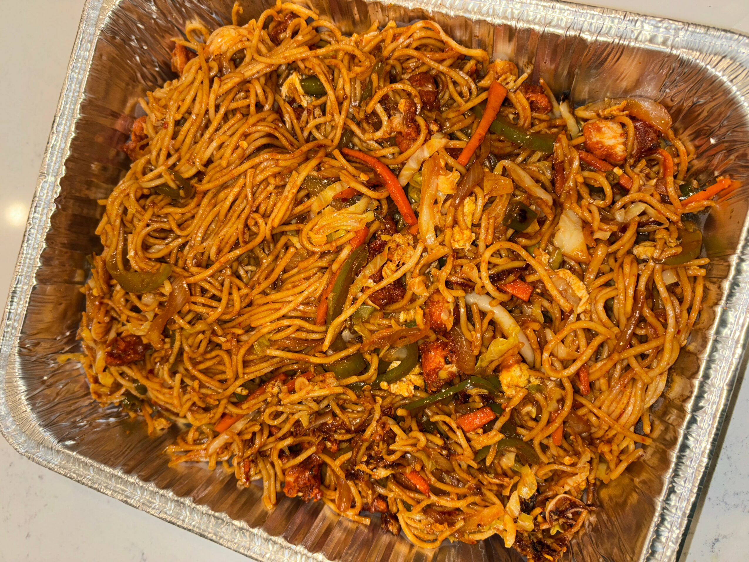 Noodles with vegetables in a foil container, including carrots and green peppers, mixed with sauce.