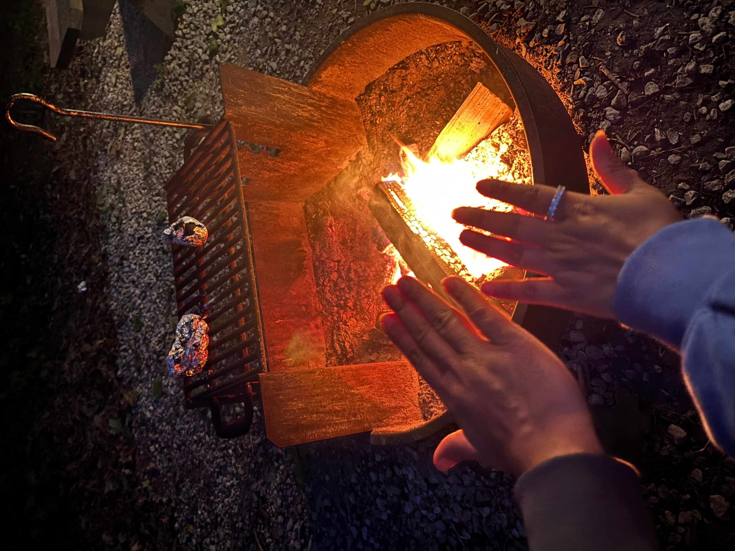 Person's hands near a small campfire on a wooden surface outdoors, with a grill and gravel ground visible.