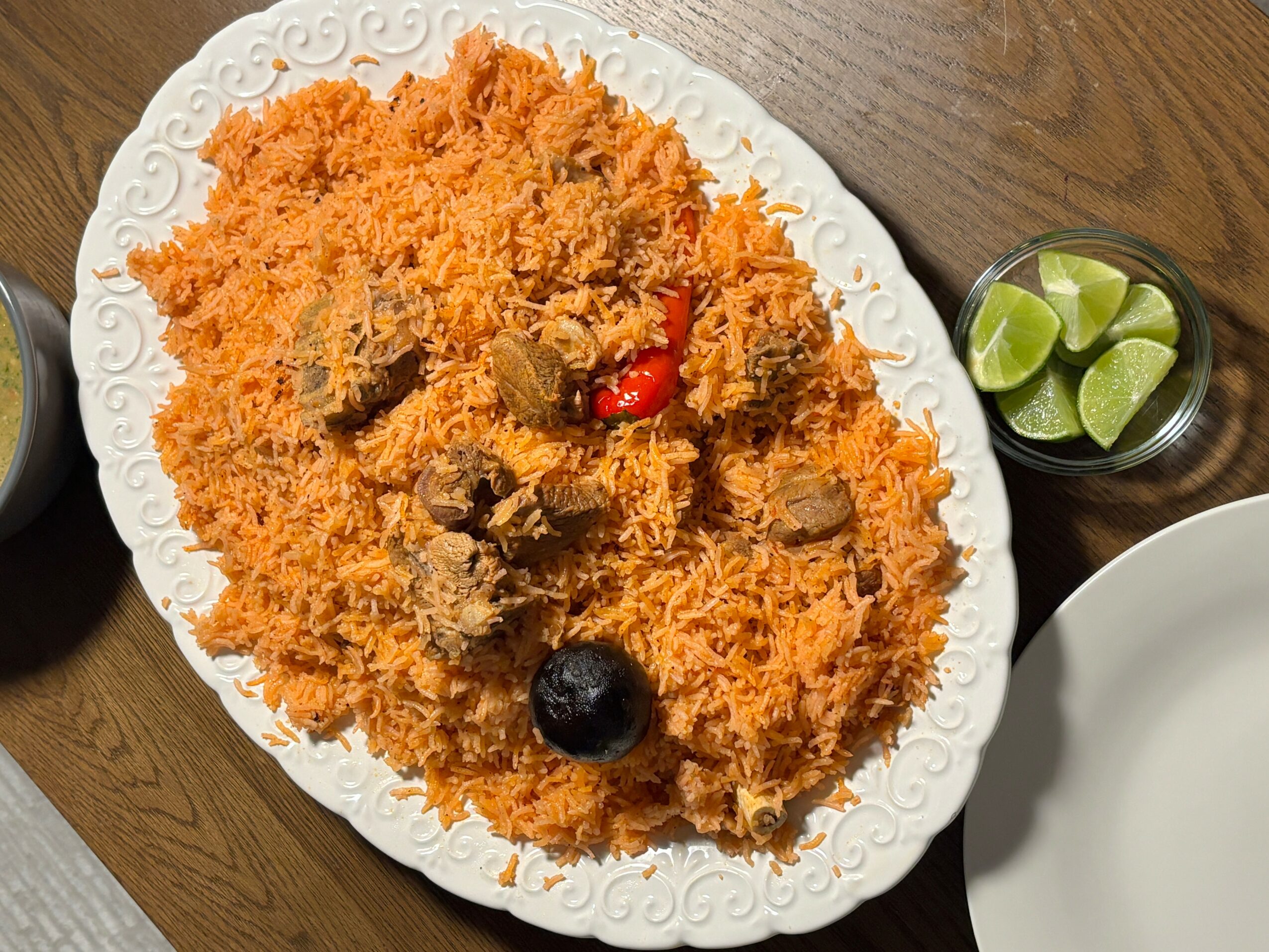Plate of fried rice with pieces of fried chicken, cherry tomato, and black olive, on a wooden table with lime wedges nearby.