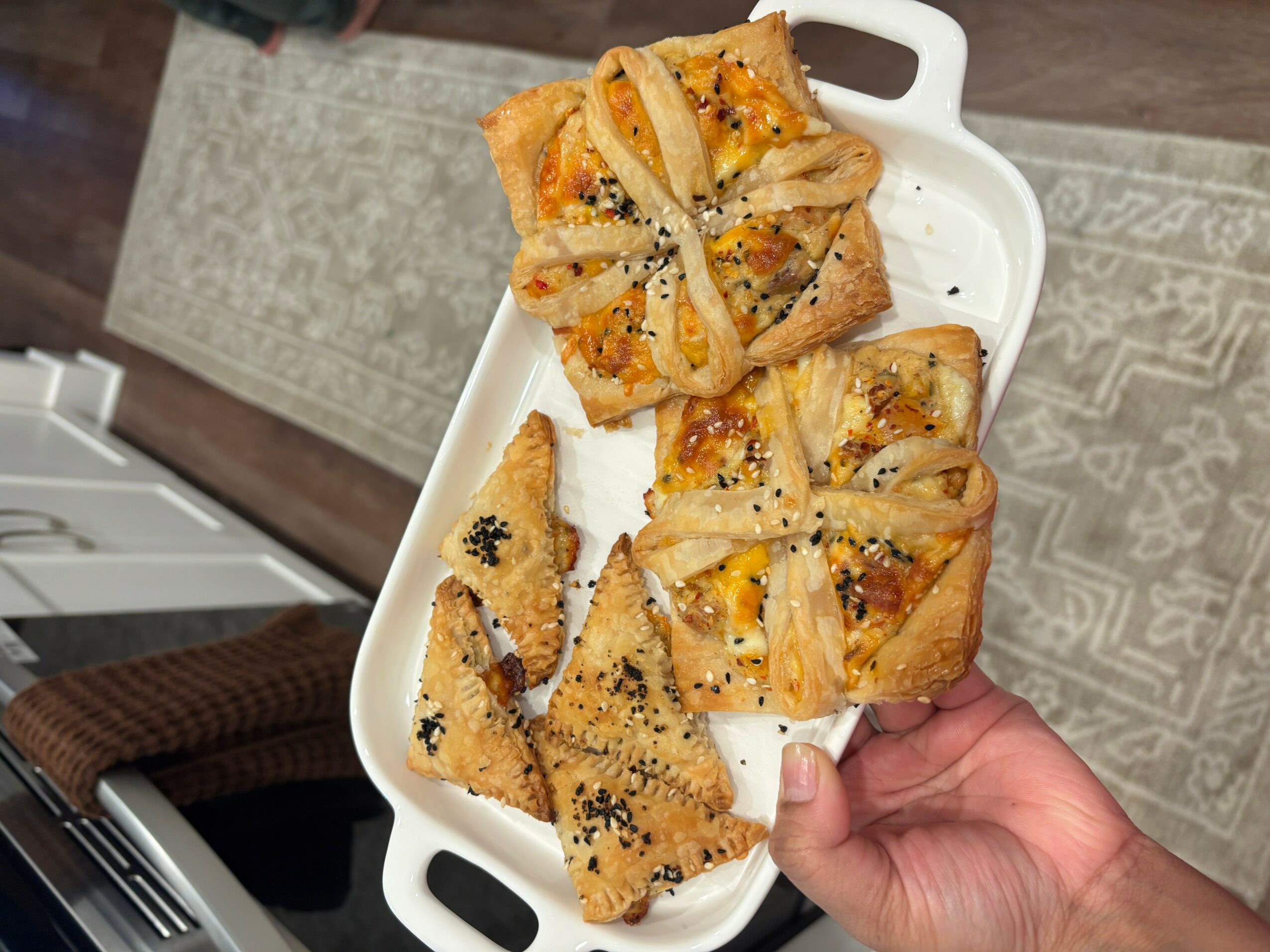 Hand holding a white tray with assorted baked pastries, some topped with sesame seeds, on a patterned rug.