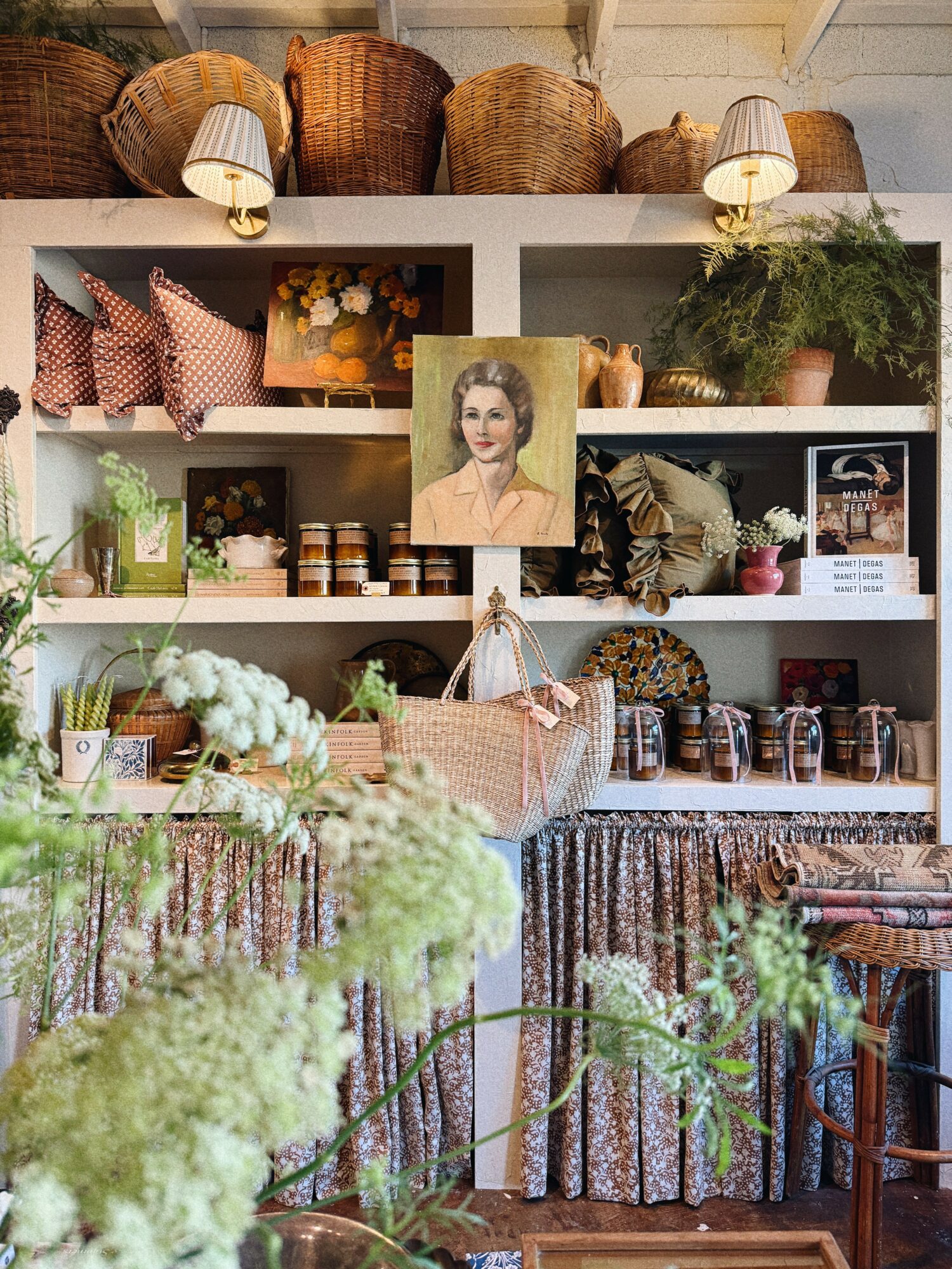 Shelves with baskets, pillows, plants, and a portrait, surrounded by flowers and greenery in a cozy shop setting.