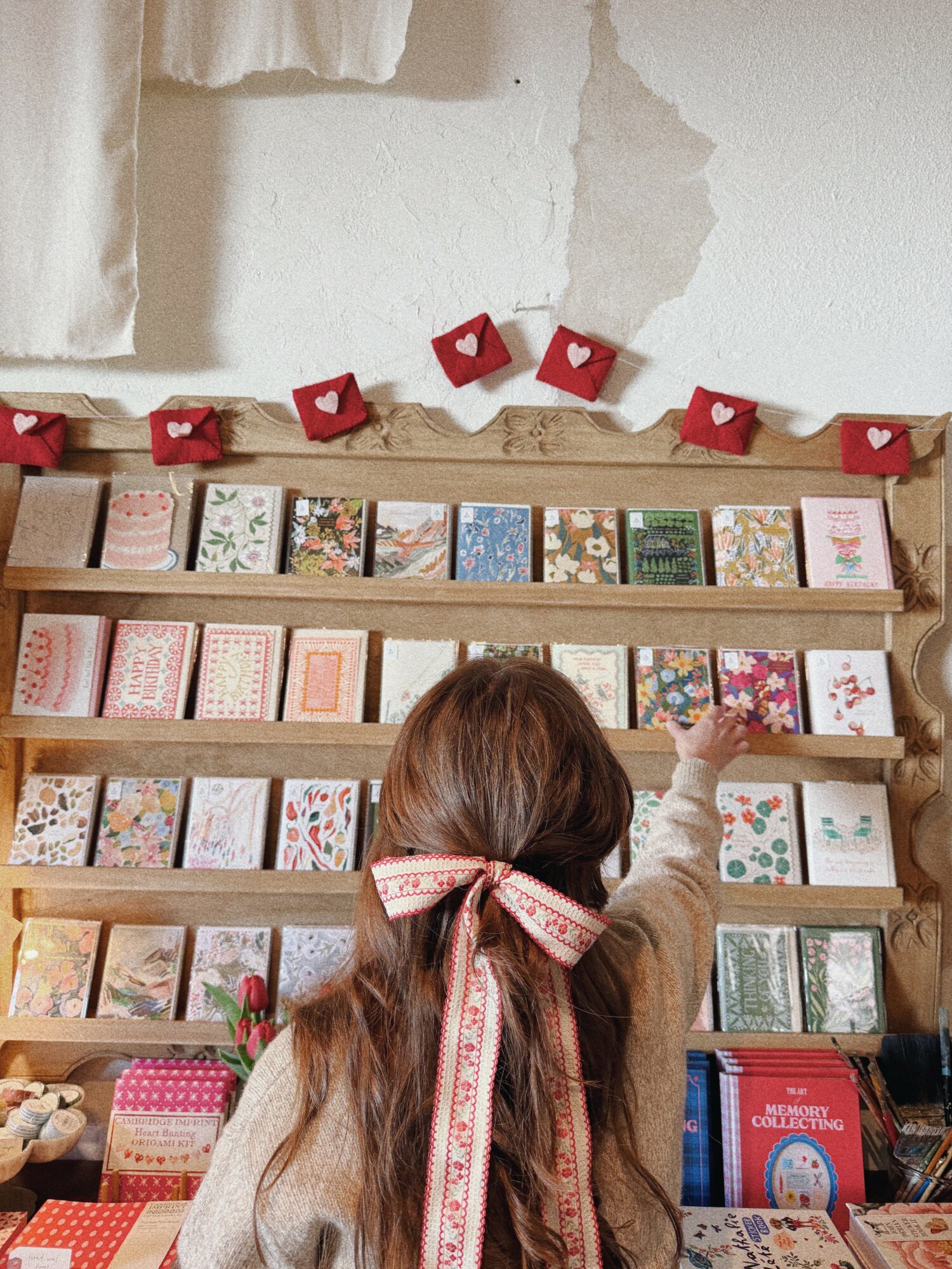 Child with long hair and a pink ribbon points at a book on a wooden bookshelf, with a torn wall above.