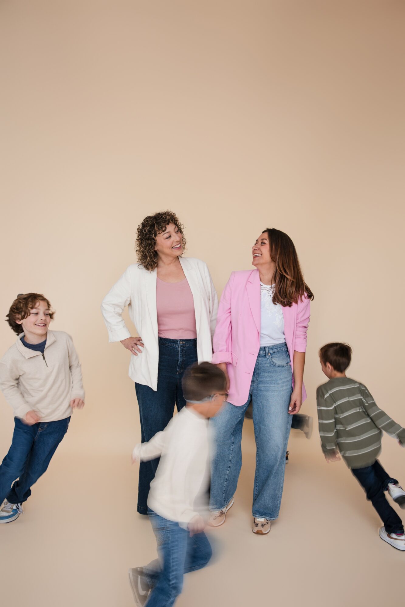 Two women stand together smiling, surrounded by four children playing around them, against a plain beige background.