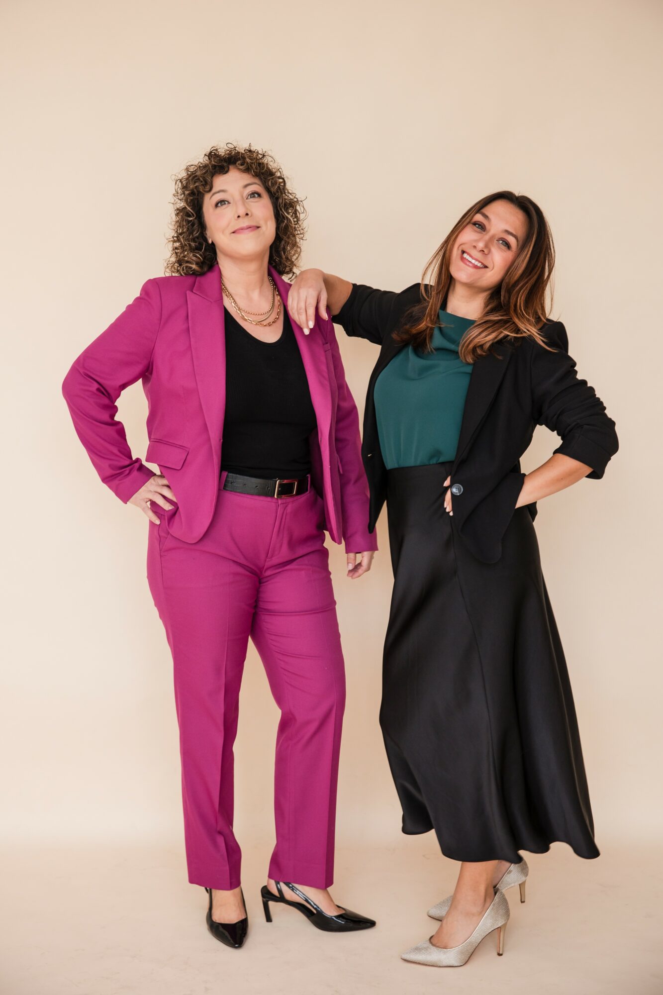 Two women stand together, one in a pink suit and the other in a black skirt and teal top, smiling.