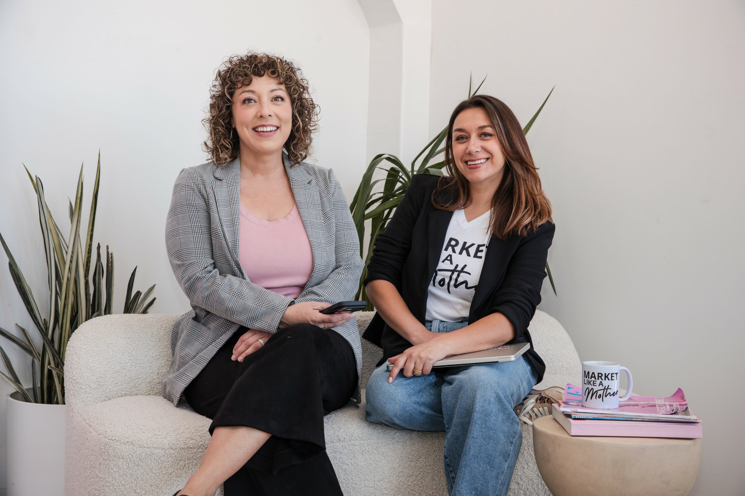 Two women sitting on a white sofa, smiling, with plants and magazines nearby, in a bright room.