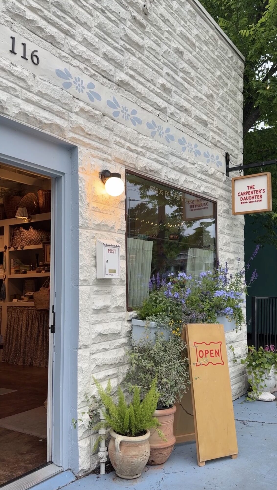 Store entrance with potted plants, a sign reading 'OPEN,' and a brick wall with a window and light fixture.