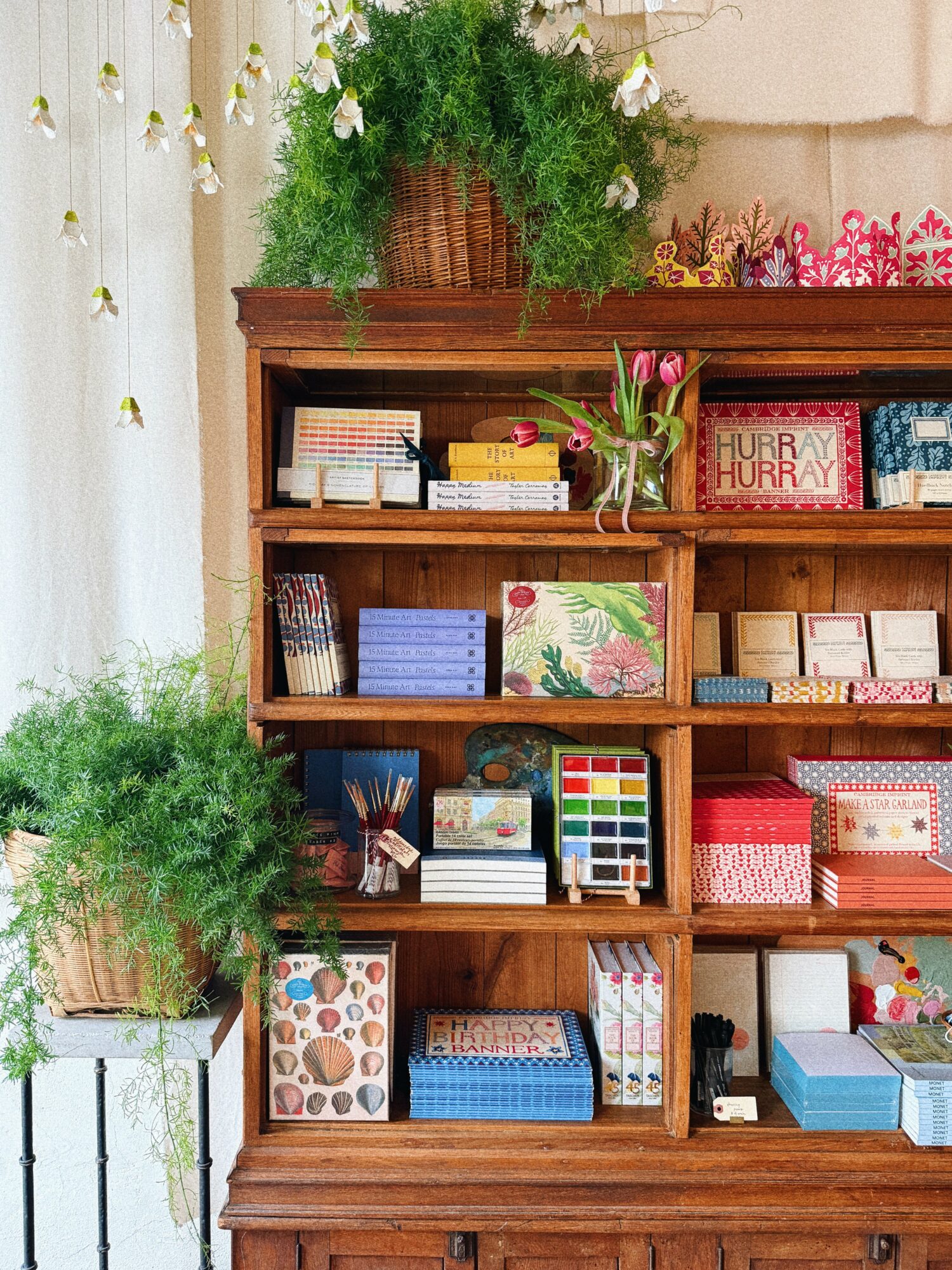 Wooden bookshelf with books, a plant, and decorative items, against a light-colored wall with curtains, and a potted plant nearby.